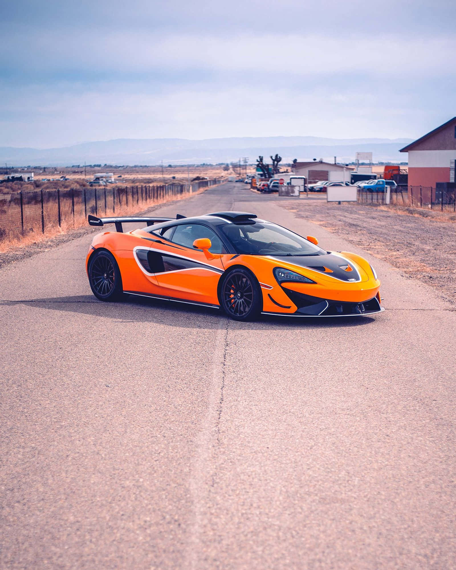 Mclaren 620r 桌布 壁紙 — 一輛充滿活力的橙色McLaren跑車，帶有黑色飾邊，停泊在鄉村瀝青路上，天空多雲。