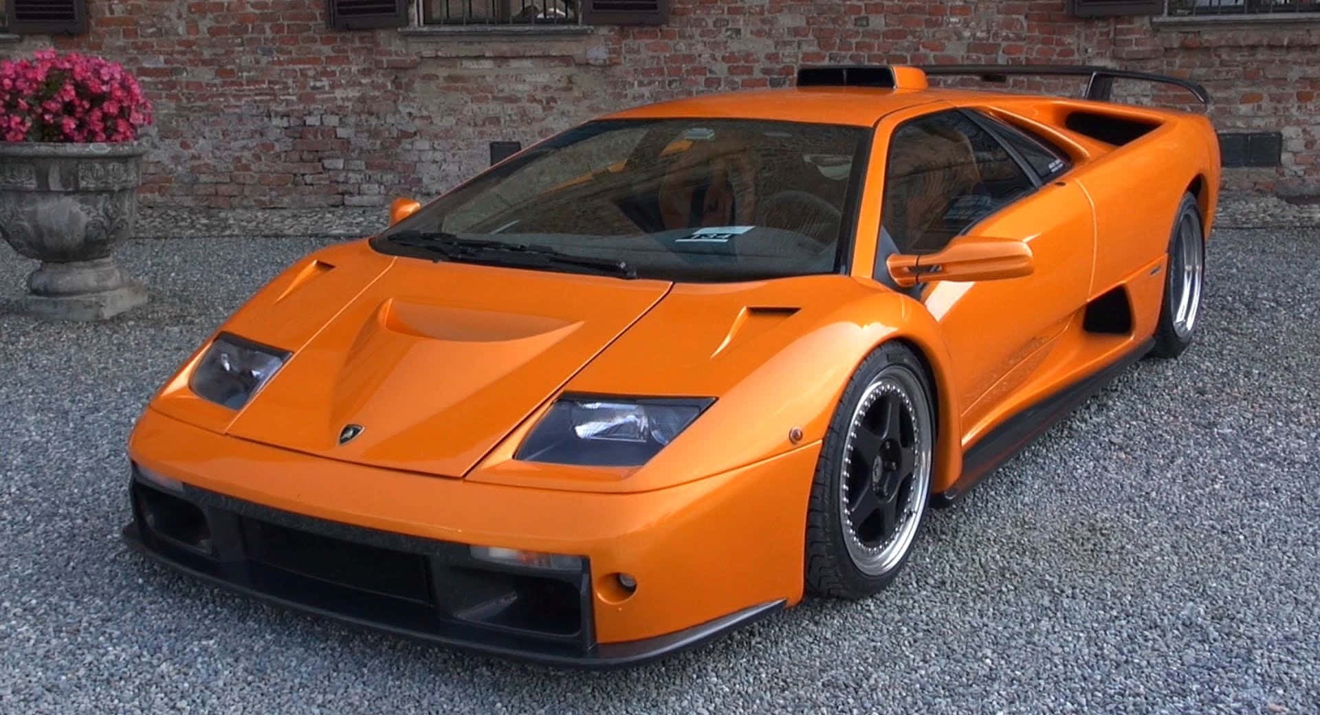 Lamborghini Diablo 桌布 壁紙 — 一輛充滿活力的橙色林寶堅尼 Diablo SV 在日光下閃閃發光，停泊在磚牆和花盆旁邊的碎石上。