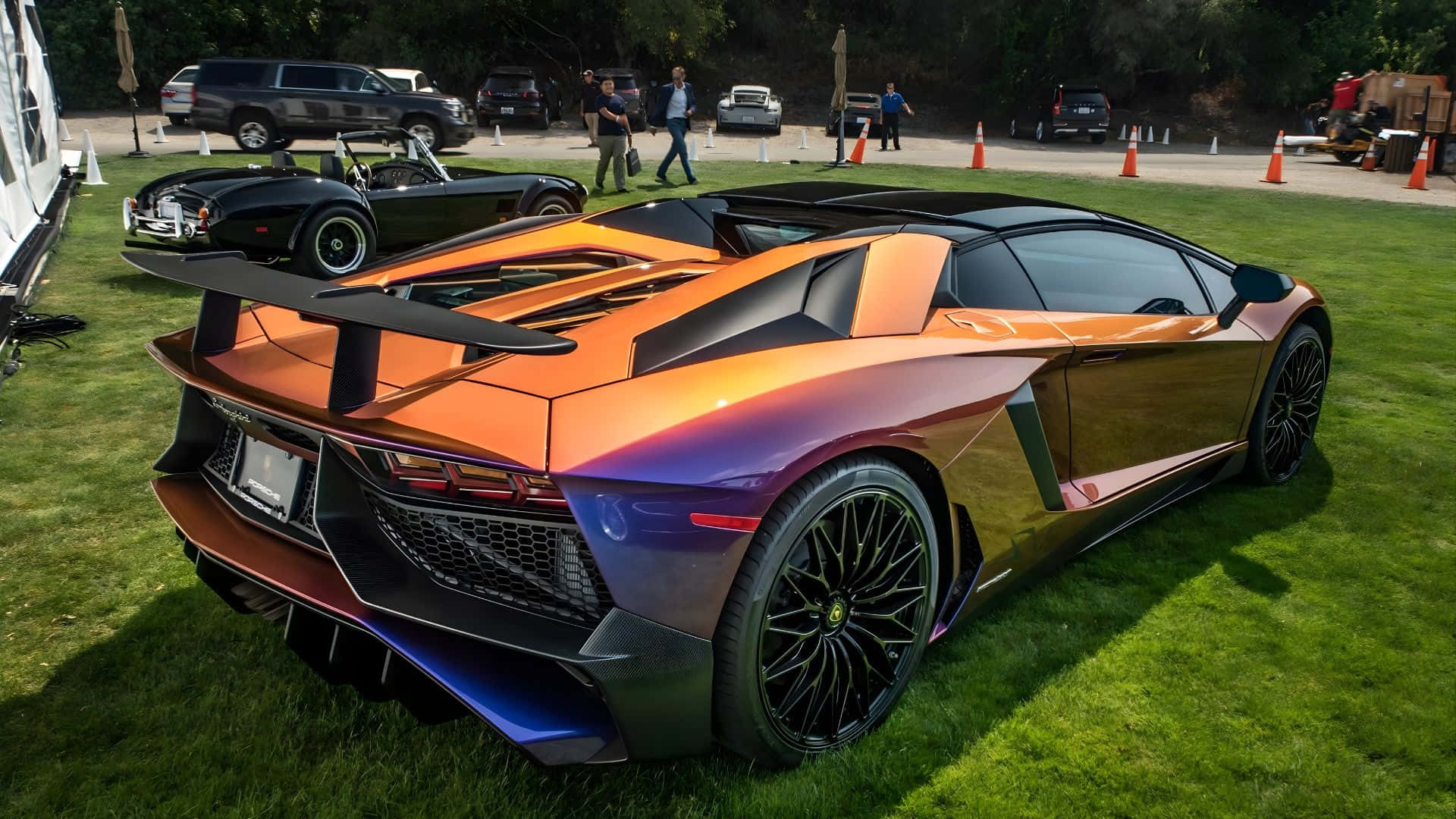  壁紙 — 一輛充滿活力的橙色和紫色林寶堅尼Aventador，帶有黑色點綴，停在車展的綠色草地上。