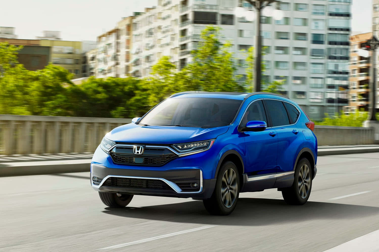Honda Cr-v 桌布 壁紙 — 一輛充滿活力的藍色本田CR-V在現代建築和茂密綠樹背景下的城市橋樑上平穩行駛。