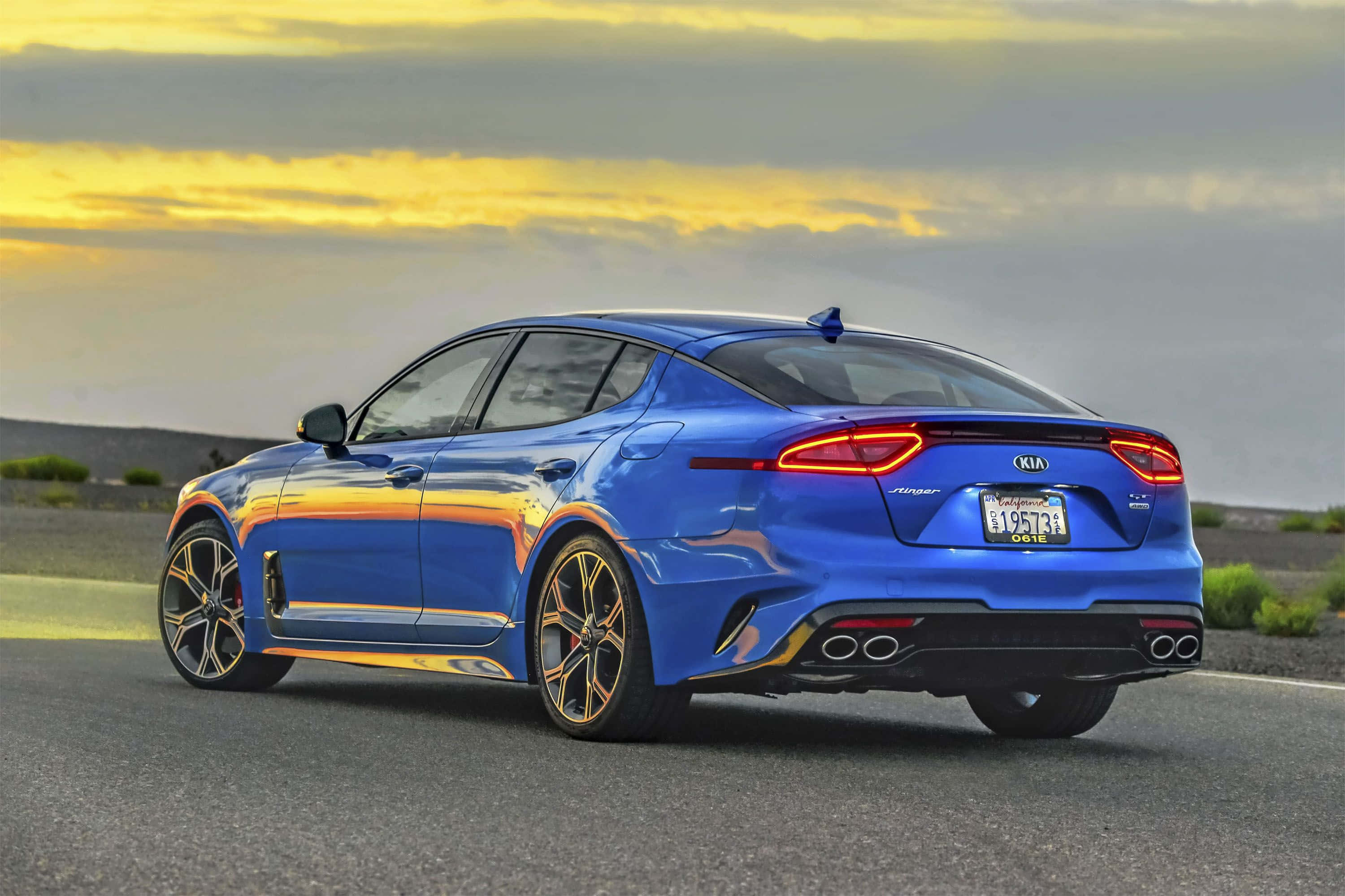 Kia Stinger 桌布 壁紙 — 一輛充滿活力的藍色Kia Stinger運動轎車在沙漠公路上閃耀，夕陽下金色雲彩映照在其流線型車身上。