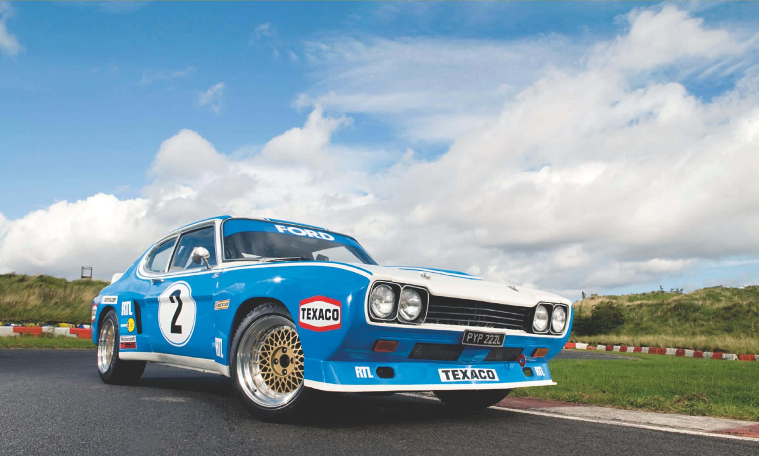Ford Escort 桌布 壁紙 — 一輛帶有Texaco標誌的藍白色福特賽車，在明亮多雲的天空下沿著蜿蜒的瀝青賽道飛馳。