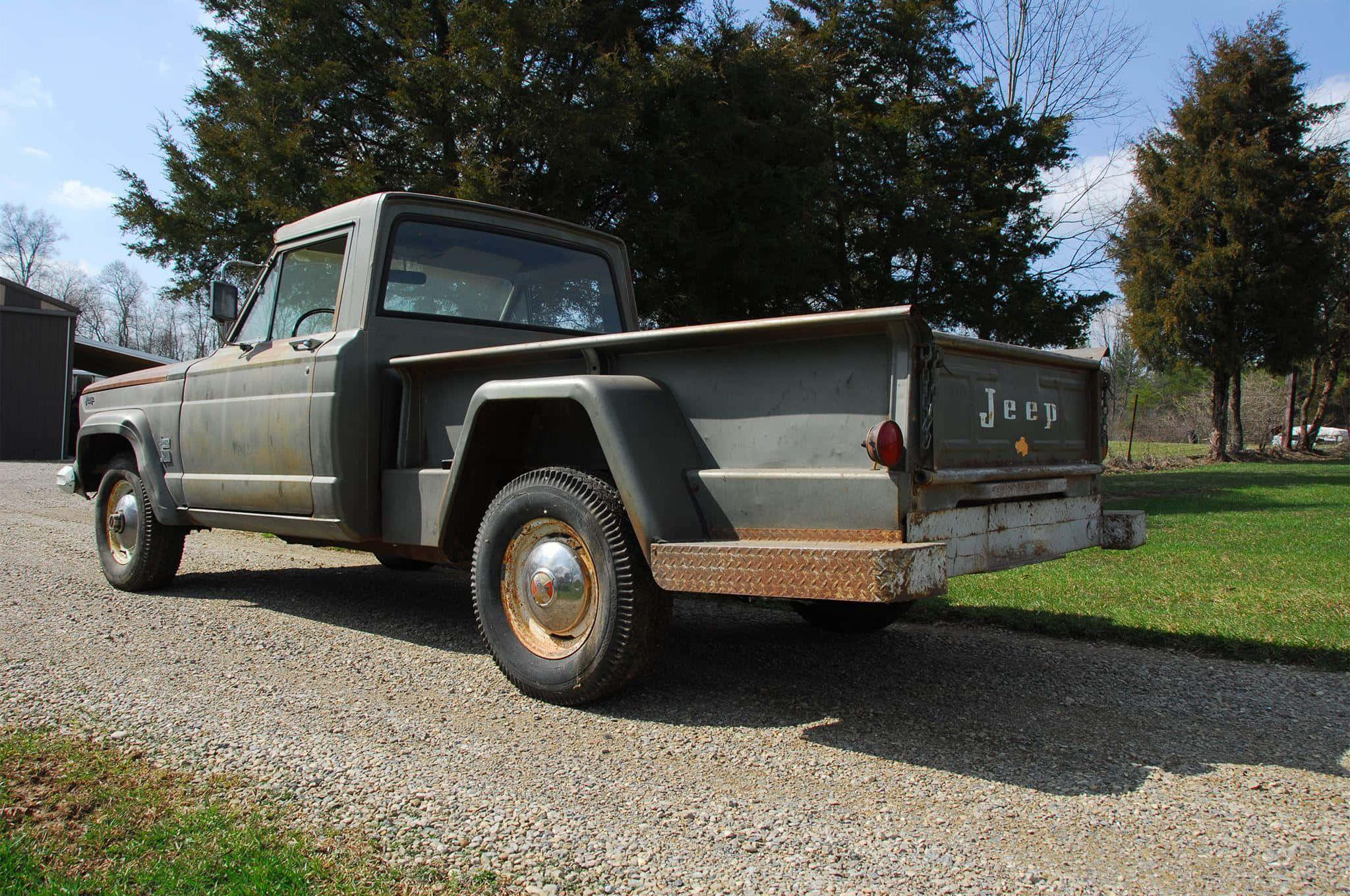 Jeep Gladiator 桌布 壁紙 — 一輛風化的灰色Jeep皮卡車停泊在碎石車道上，周圍是高聳的常綠樹木，天空清澈湛藍。