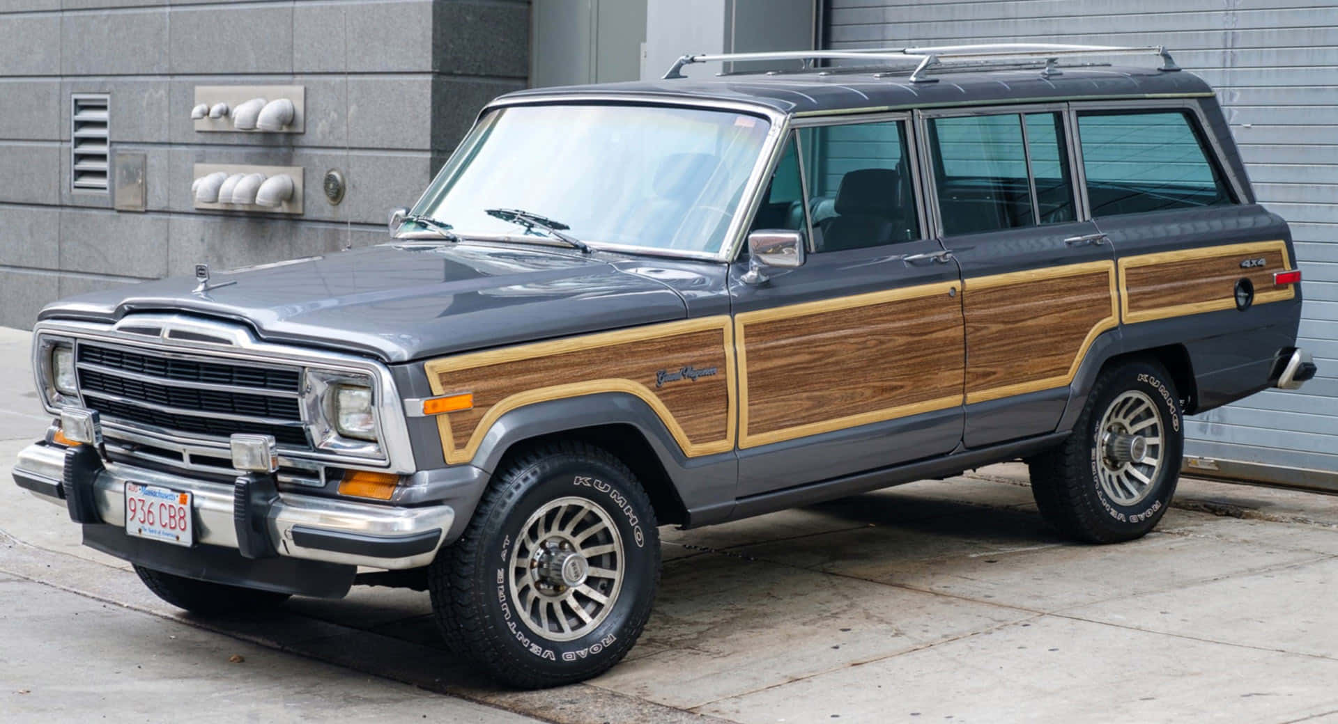 Jeep Wagoneer 桌布 壁紙 — 一輛經典灰色Jeep Wagoneer配備木紋飾板，停在灰色磚砌建築旁的混凝土行人路上。