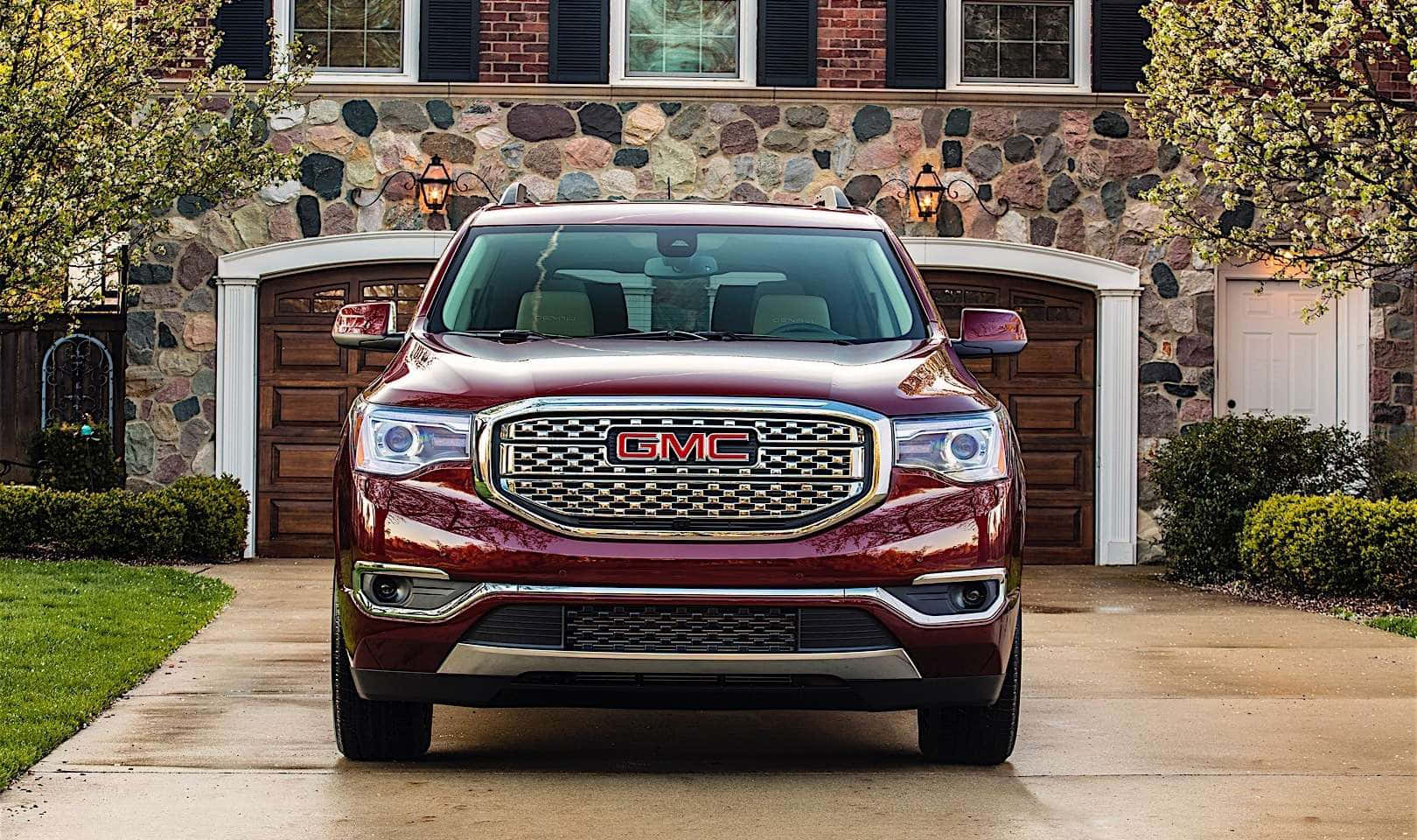 Gmc Acadia 桌布 壁紙 — 一輛光亮的棗紅色GMC Acadia SUV停泊在石面房屋前的混凝土車道上，房屋有木製車庫門和綠色灌木。