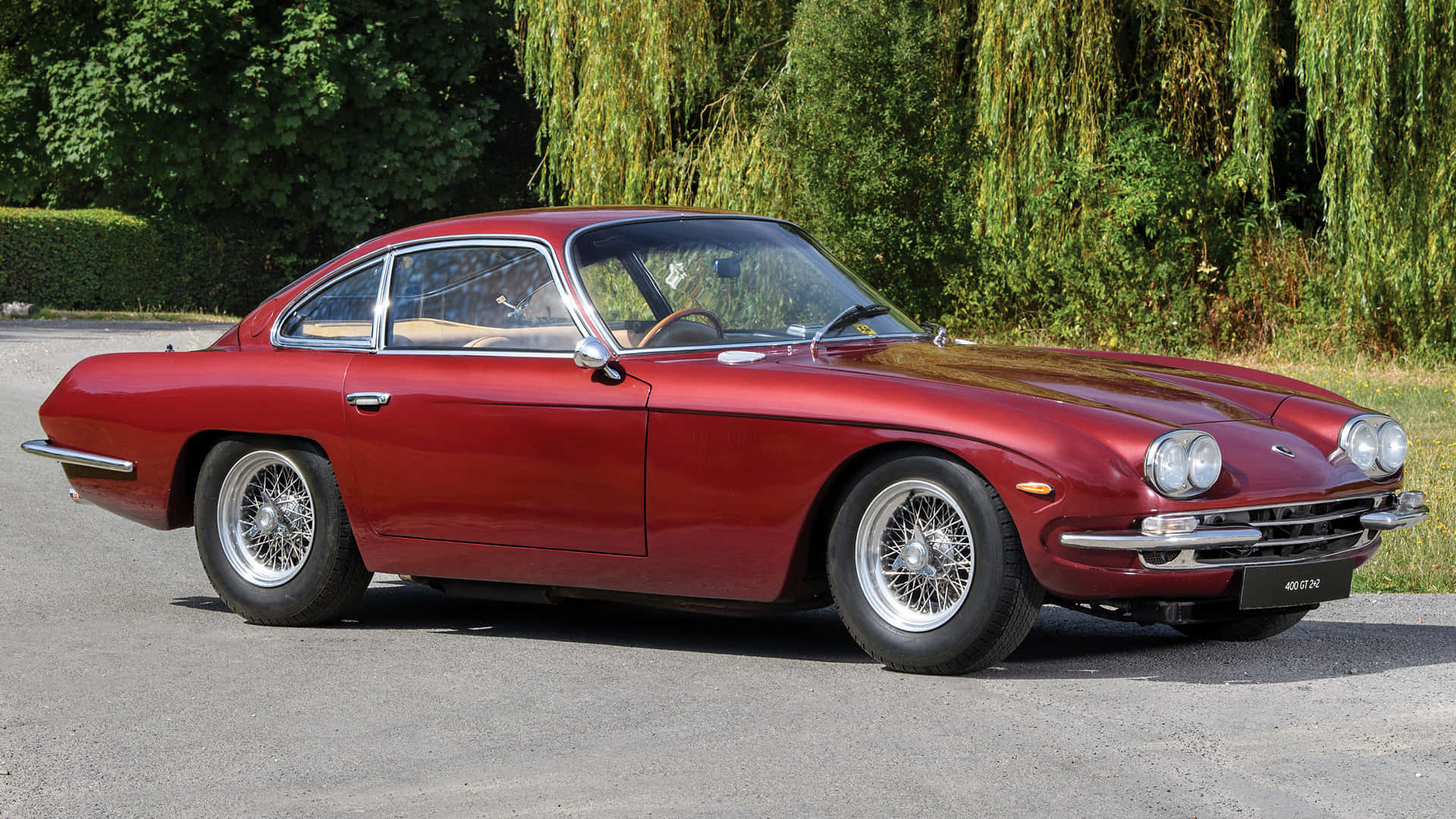 Lamborghini 400 Gt 桌布 壁紙 — 一輛光亮的紅色經典跑車，配備鋼絲輪圈，停在瀝青路面上，背後是茂密的綠樹，在明亮的日光下。