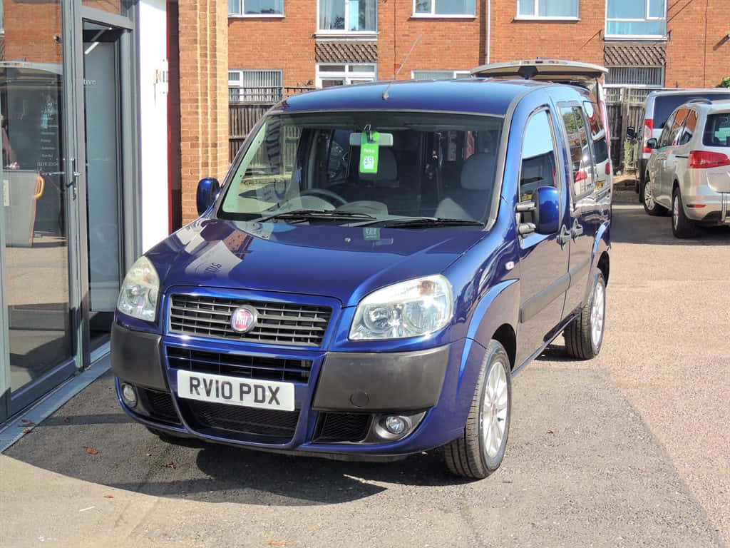  壁紙 — 一輛光亮的藍色Fiat Panda 4x4停泊在鋪砌的停車場，旁邊是帶玻璃門的磚砌建築。
