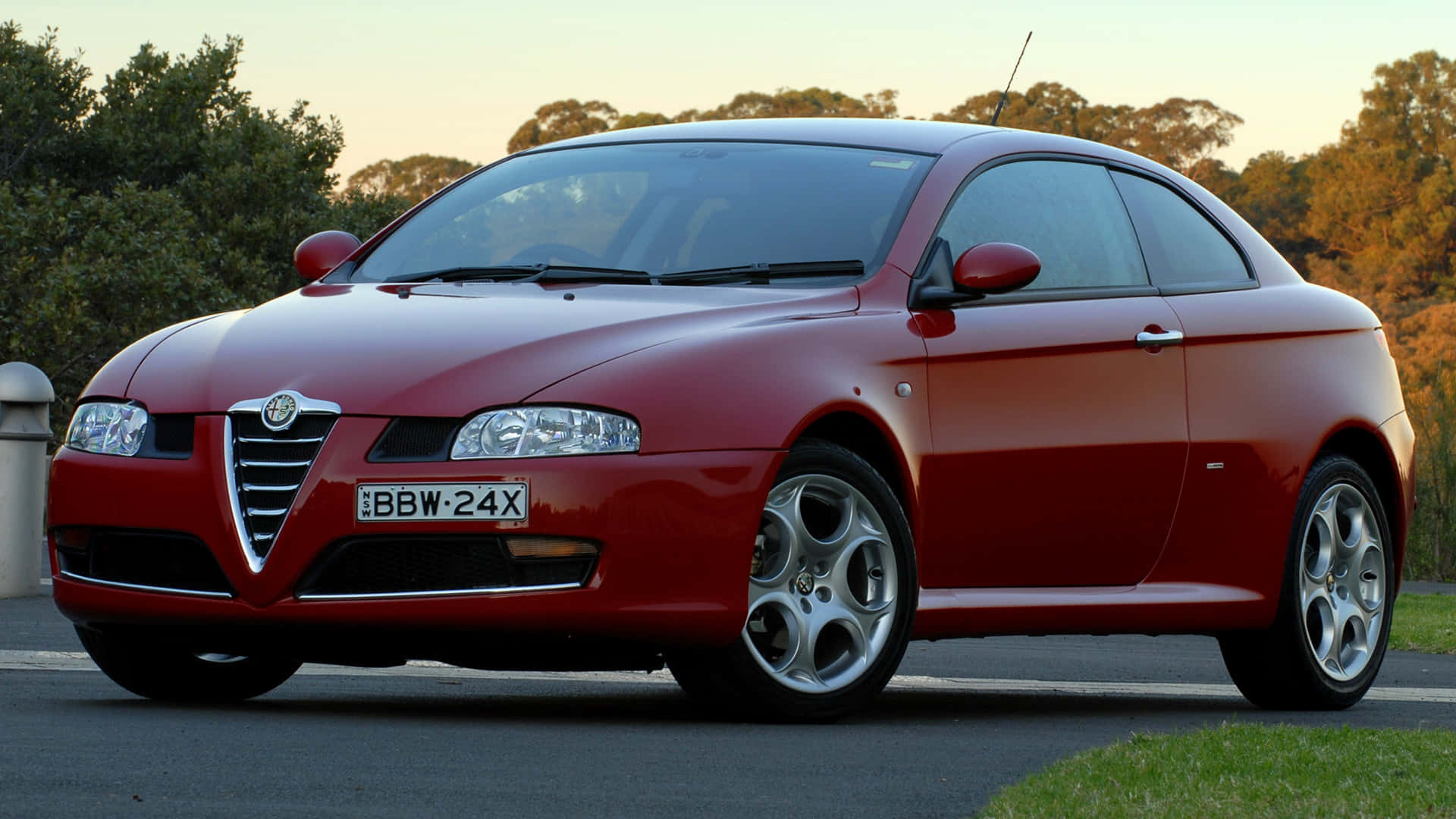 Alfa Romeo Gt 桌布 壁紙 — 一輛亮紅色嘅Alfa Romeo雙門跑車喺黃金時段停泊喺路邊，背景有樹木。