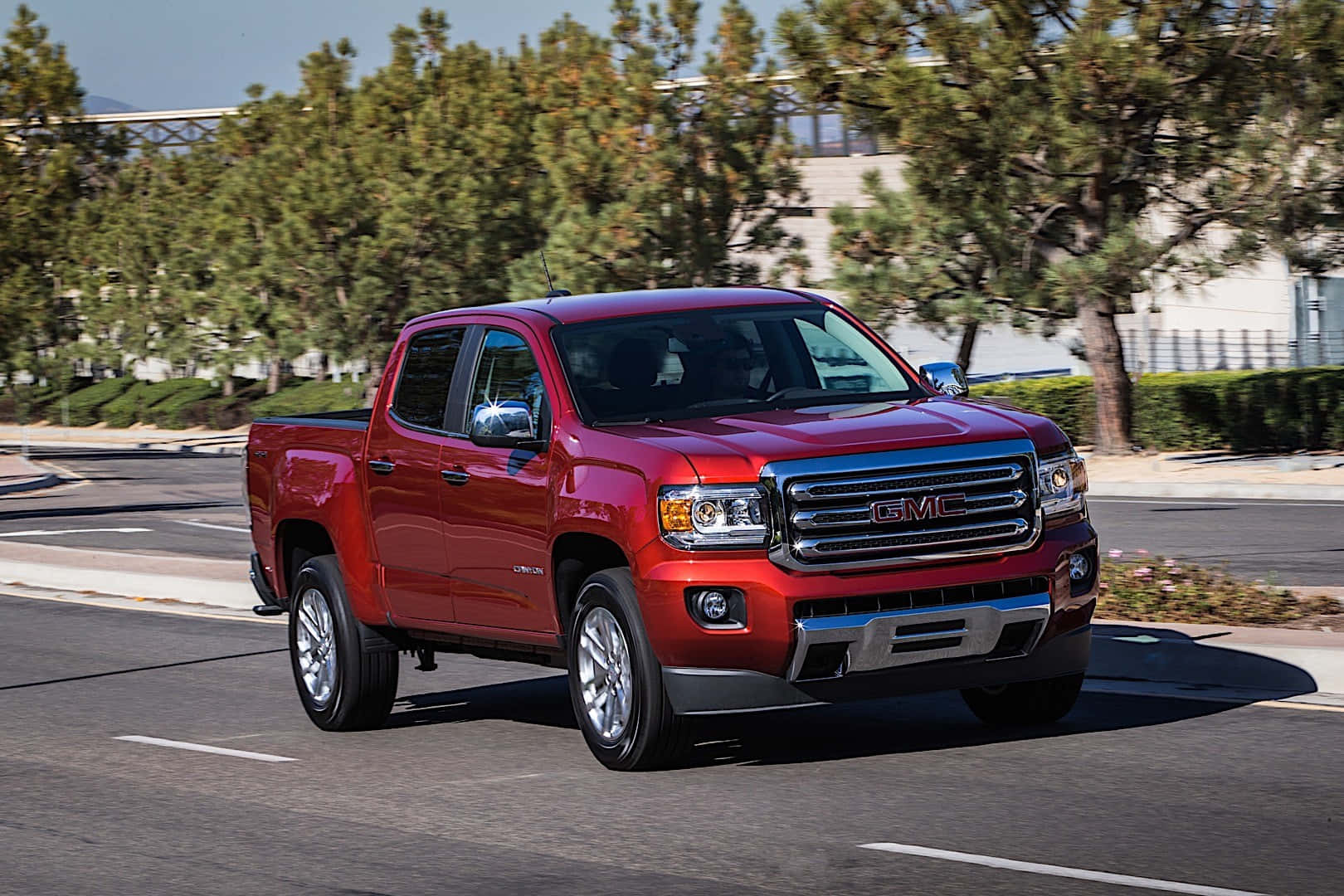 Gmc Canyon 桌布 壁紙 — 一輛亮紅色的GMC Canyon皮卡車在陽光明媚的道路上行駛，道路兩旁綠樹成蔭，現代建築林立。