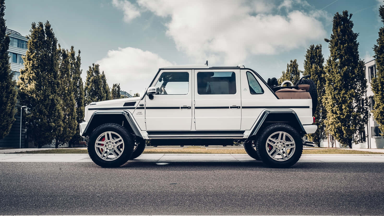  壁紙 — 一輛純白色的Mercedes-Benz G-Class SUV停泊在種滿高大常綠樹的街道上，天空局部多雲。