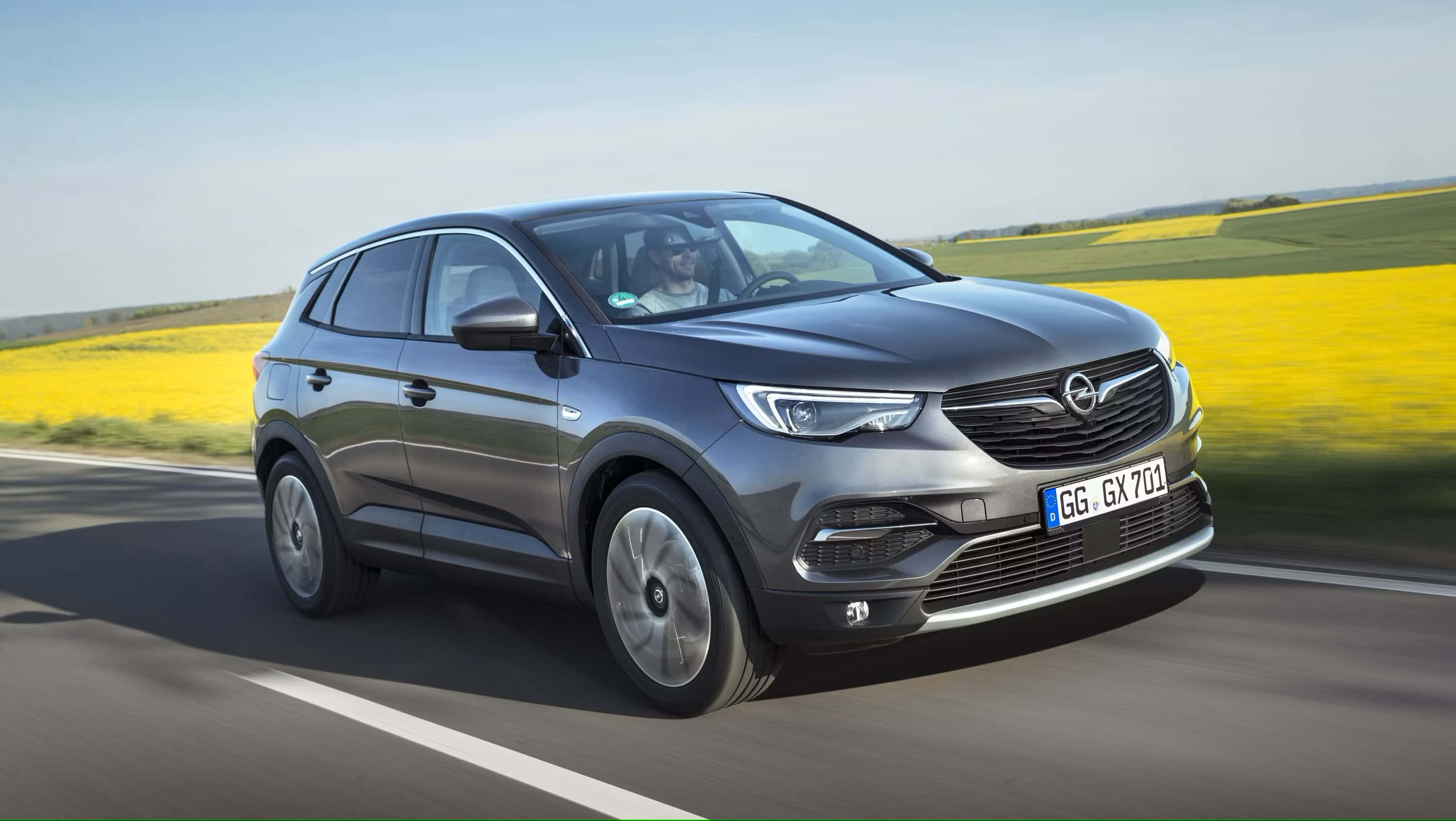 Opel Grandland X 桌布 壁紙 — 一輛時尚的灰色SUV在陽光明媚的道路上行駛，兩旁是充滿活力的黃色油菜花田，頭頂是清澈的藍天。