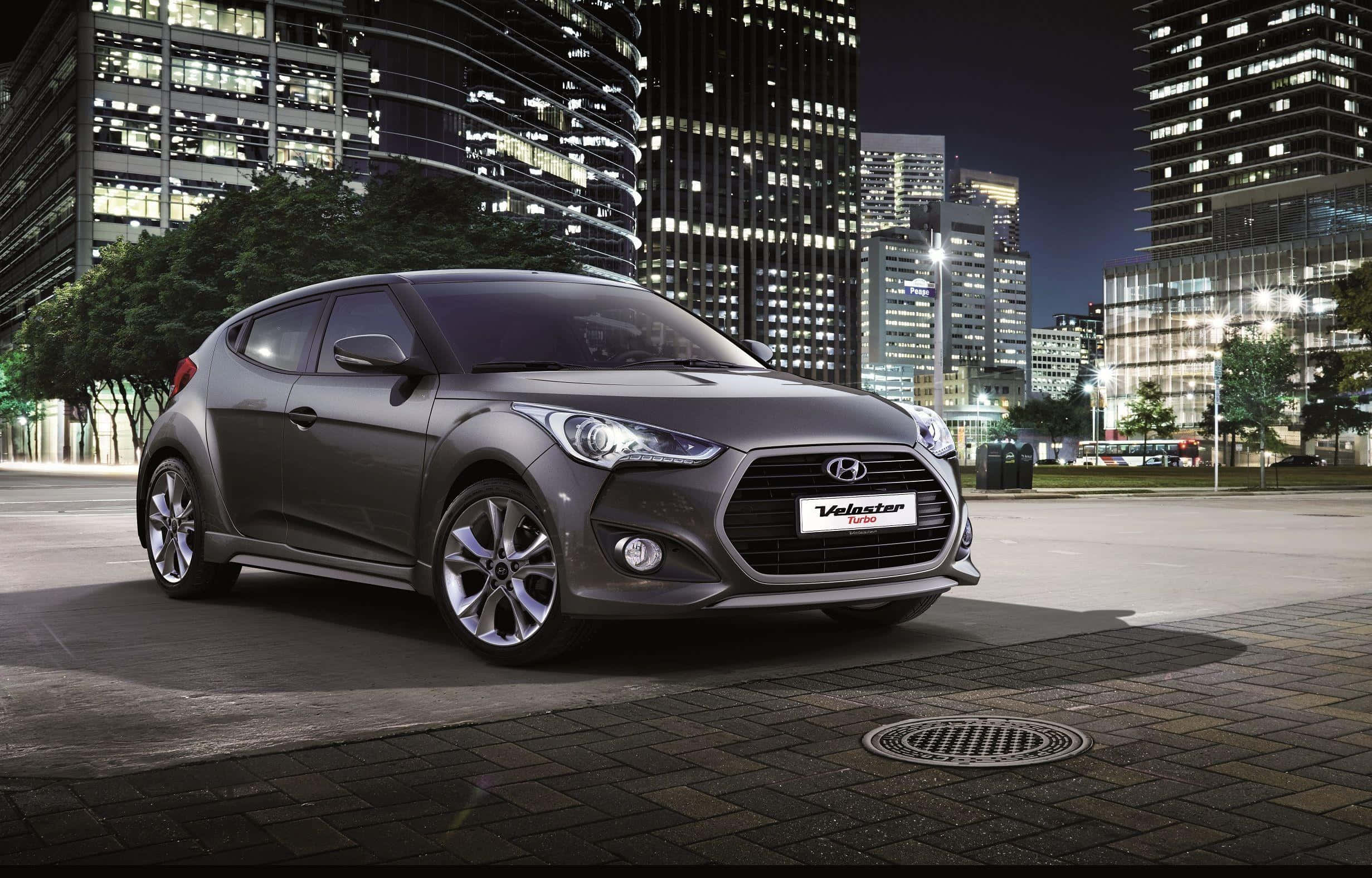 Hyundai Veloster 桌布 壁紙 — 一輛時尚的灰色現代Veloster停泊在鵝卵石街道上，夜晚現代城市天際線的燈光閃爍。
