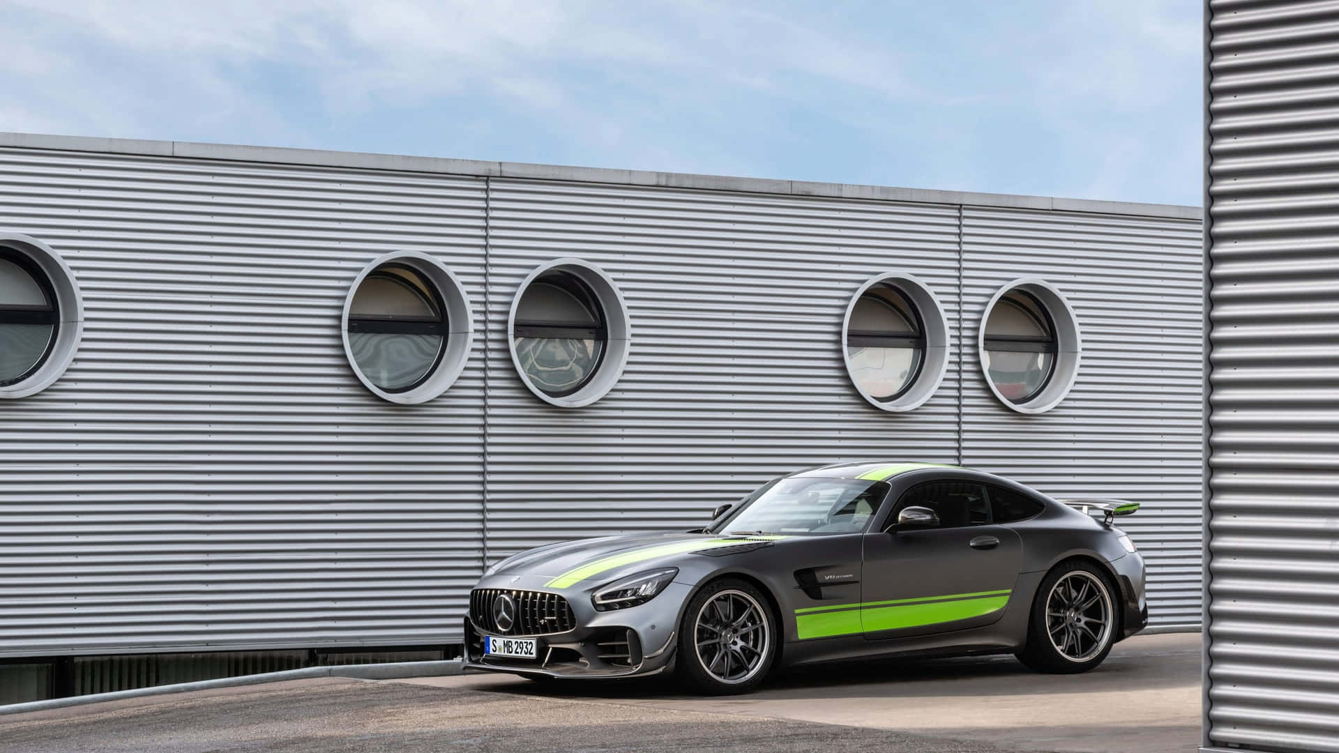  壁紙 — 一輛時尚的灰色 Mercedes-AMG GT 帶有鮮綠色點綴，停在一個帶有圓形窗戶的現代波紋金屬建築前。