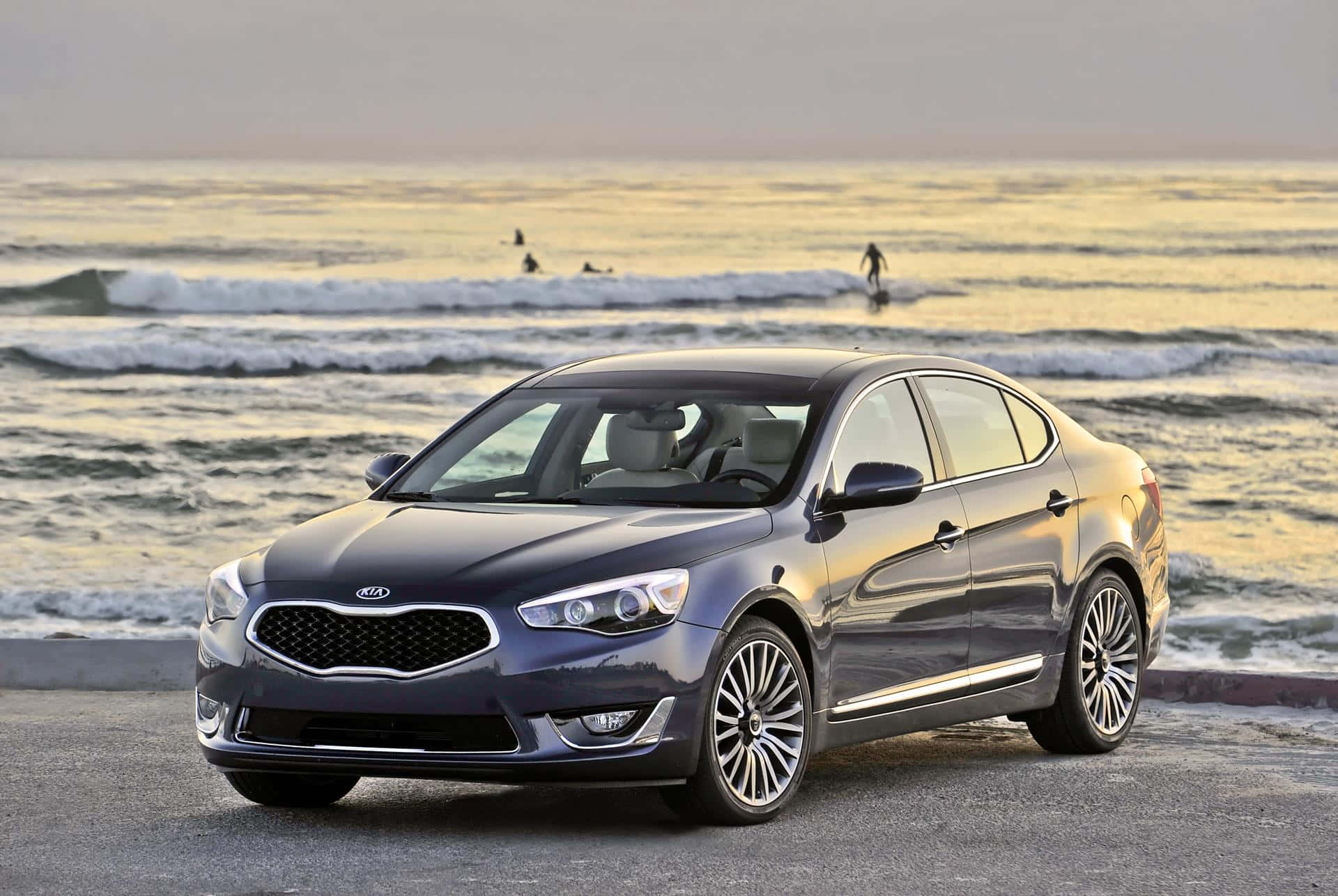 Kia Cadenza 桌布 壁紙 — 一輛時尚的深灰色Kia轎車停泊在沿海公路上，衝浪者在黃金時段乘風破浪。