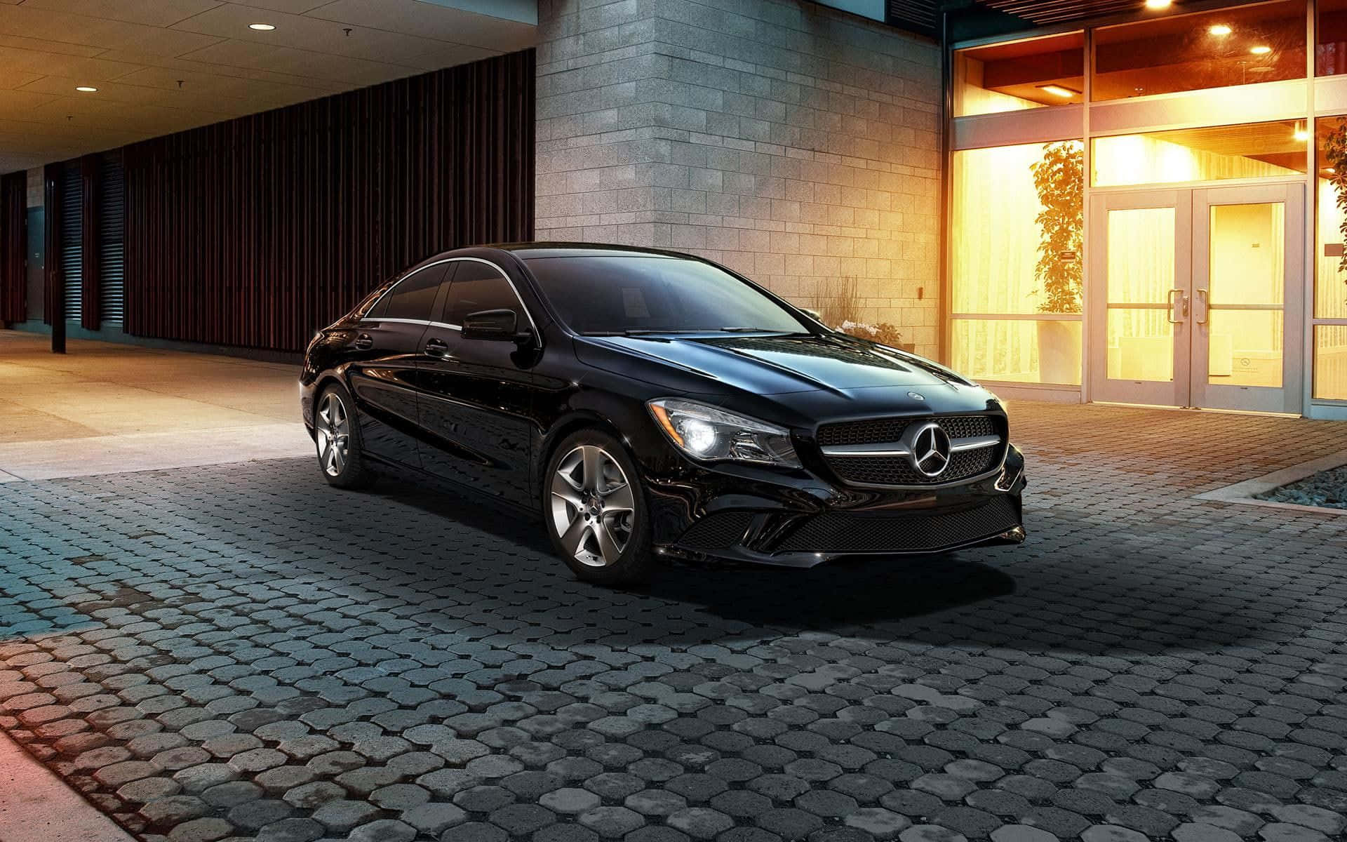 Mercedes Benz Cla-class 桌布 壁紙 — 一輛時尚的黑色平治CLA房車在溫暖的晚燈下閃閃發光，停泊在現代建築入口旁的鵝卵石車道上。