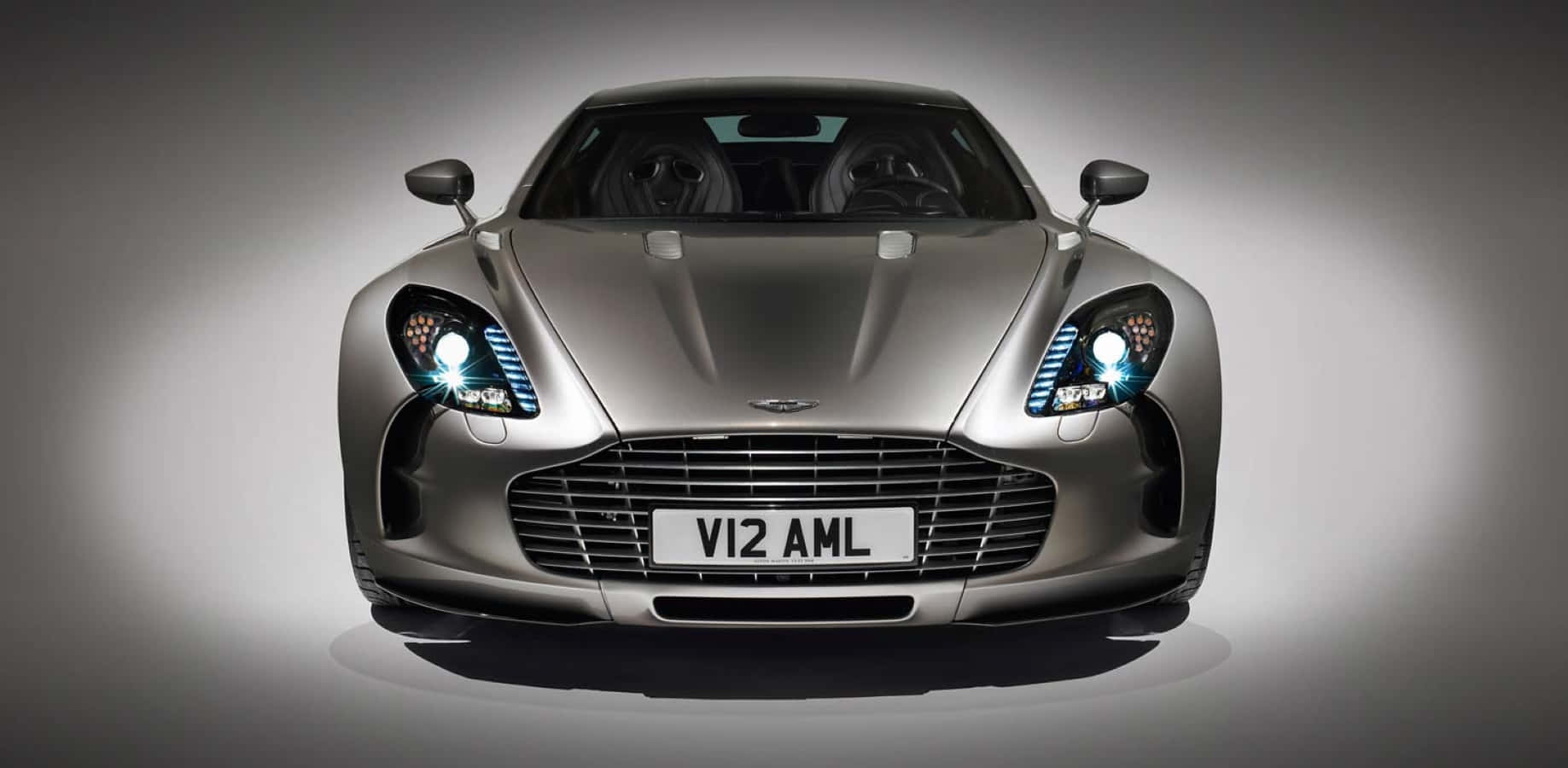 Aston Martin One-77 桌布 壁紙 — 一輛時尚的銀色Aston Martin跑車，配以發光的車頭燈，在柔和的灰色工作室背景中居中展示。