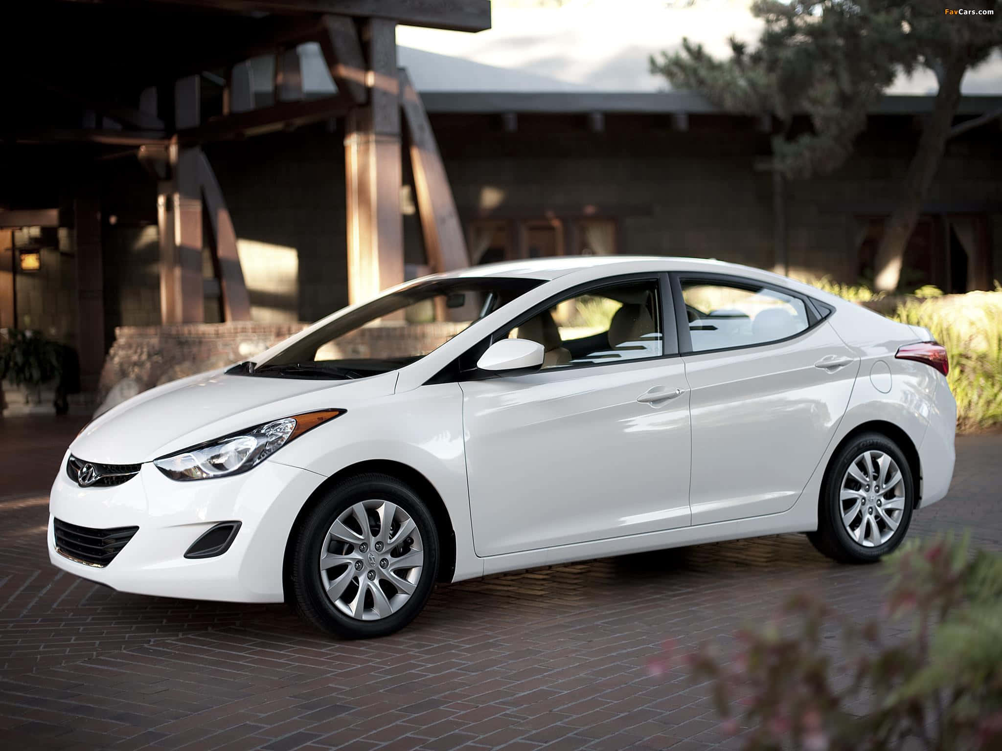 Hyundai Elantra 桌布 壁紙 — 一輛時尚的白色現代伊蘭特轎車停泊在磚砌車道上，前方是一座現代木造建築，柔和陽光投射出溫暖陰影。