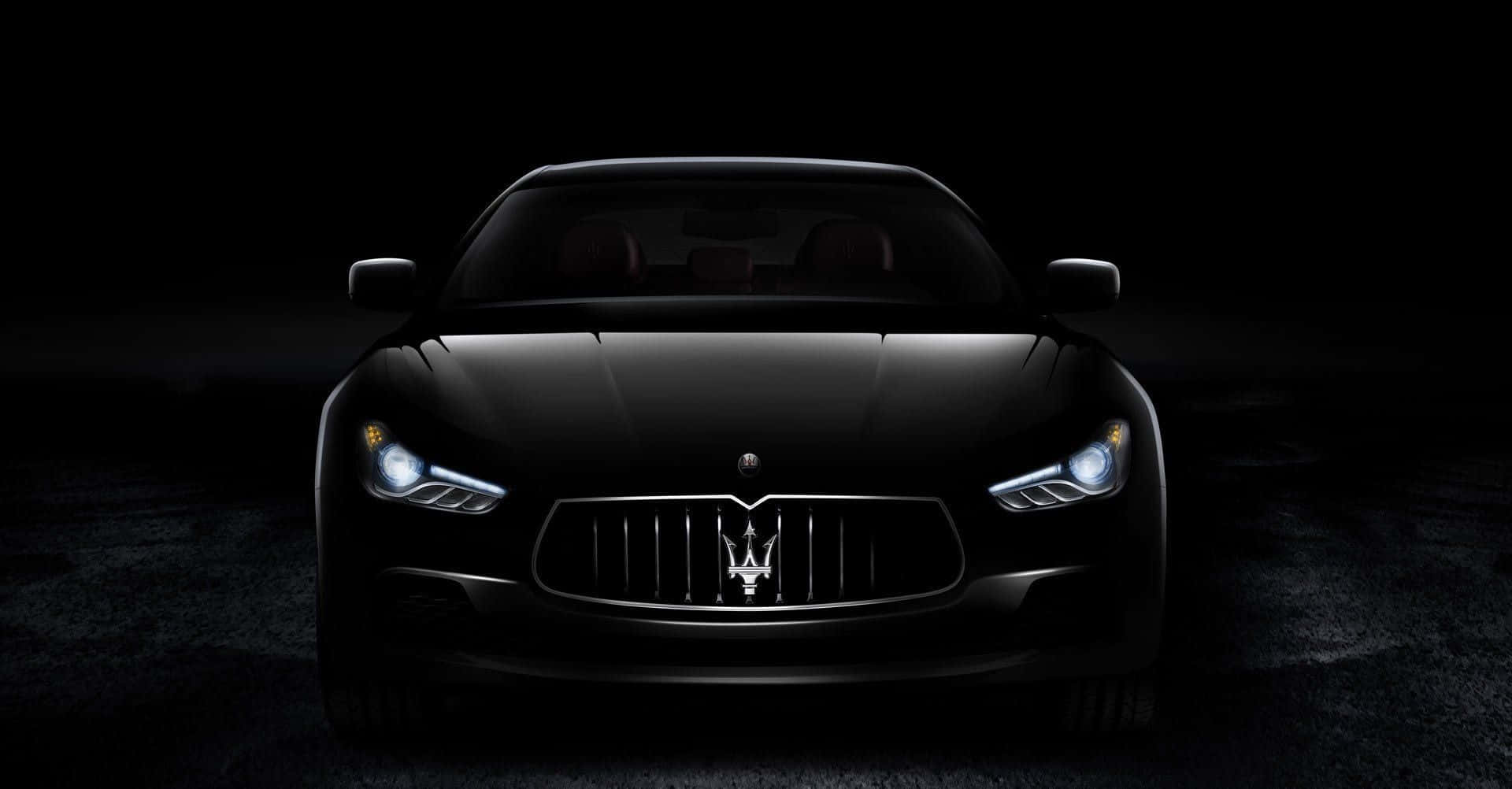 Maserati Ghibli 桌布 壁紙 — 一輛時尚的黑色瑪莎拉蒂跑車在漆黑的攝影棚背景下閃爍著銳利的車頭燈。