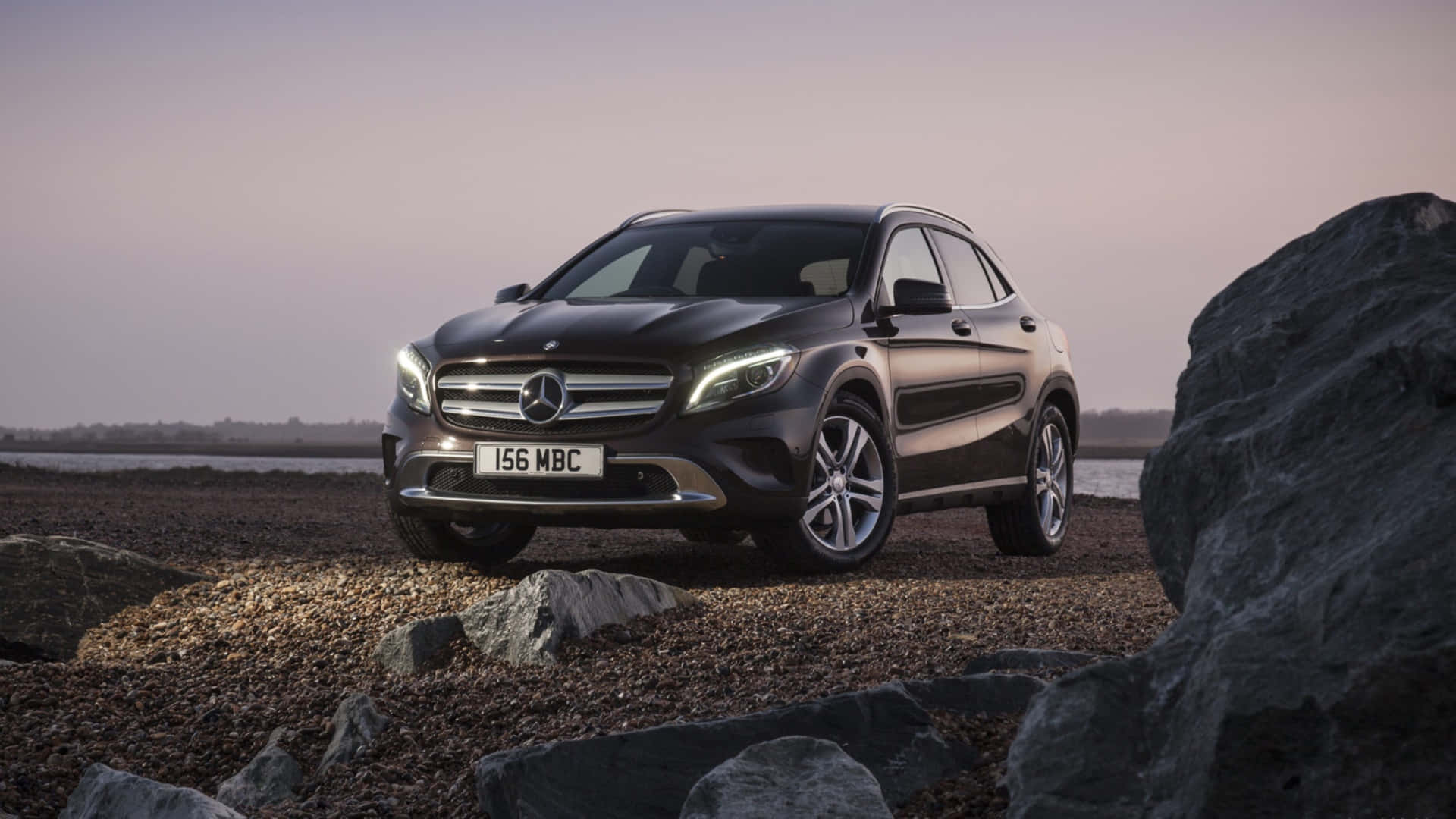 Mercedes Benz Gla-class 桌布 壁紙 — 一輛時尚的黑色Mercedes-Benz GLA-Class SUV停泊在黃昏時分的岩石海岸線上，車頭燈閃閃發光。