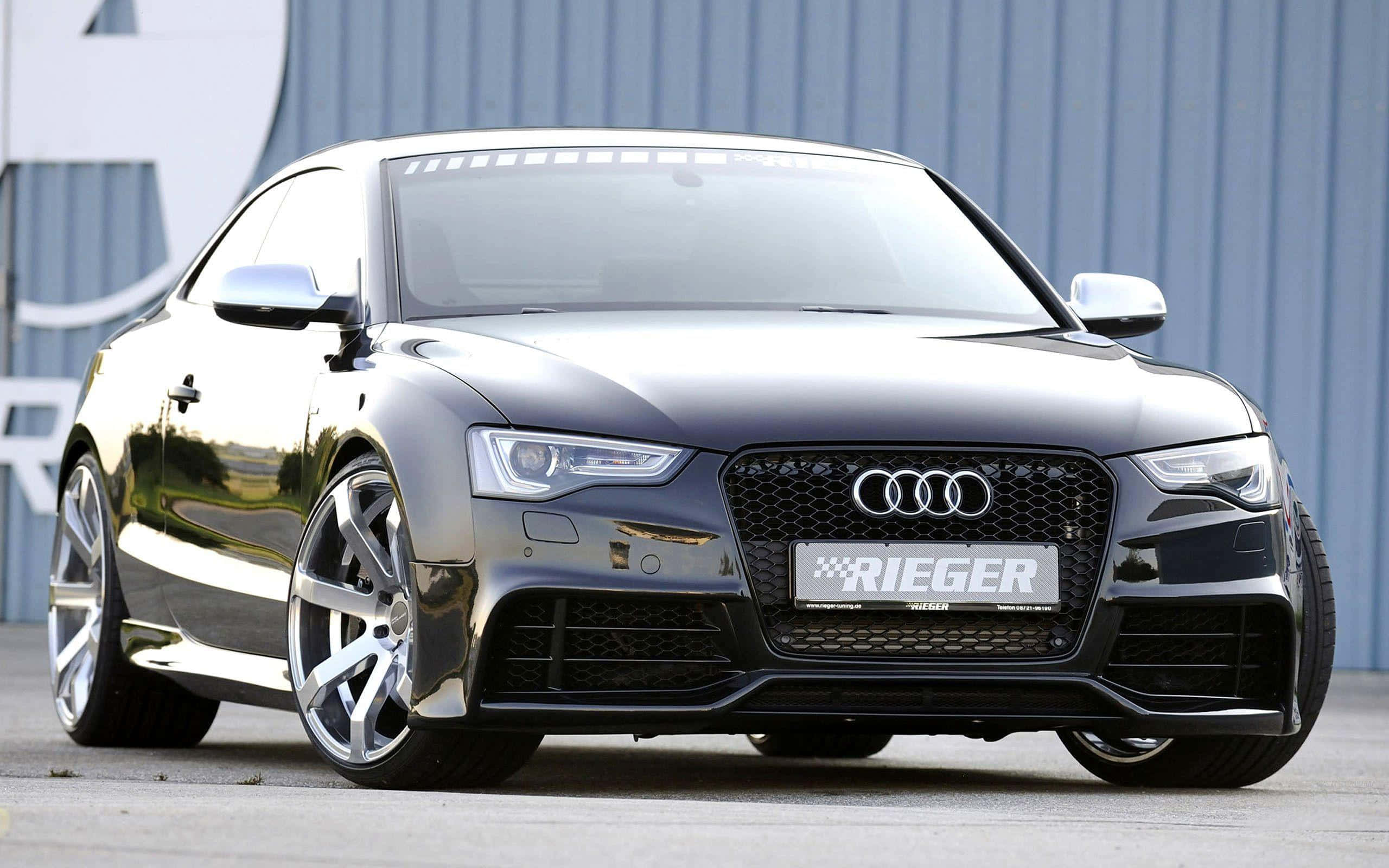 Audi A5 桌布 壁紙 — 一輛時尚的黑色奧迪RS5雙門轎跑車，配備定制Rieger輪圈，停在藍色波紋金屬建築前。