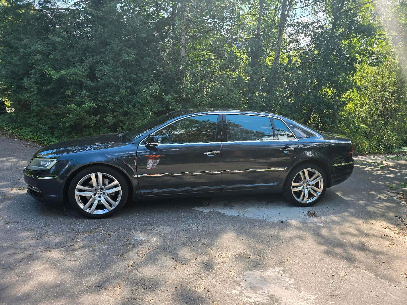  壁紙 — 一輛時尚的黑色轎車停泊在陽光斑駁的碎石車道上，周圍環繞著茂密的綠樹。