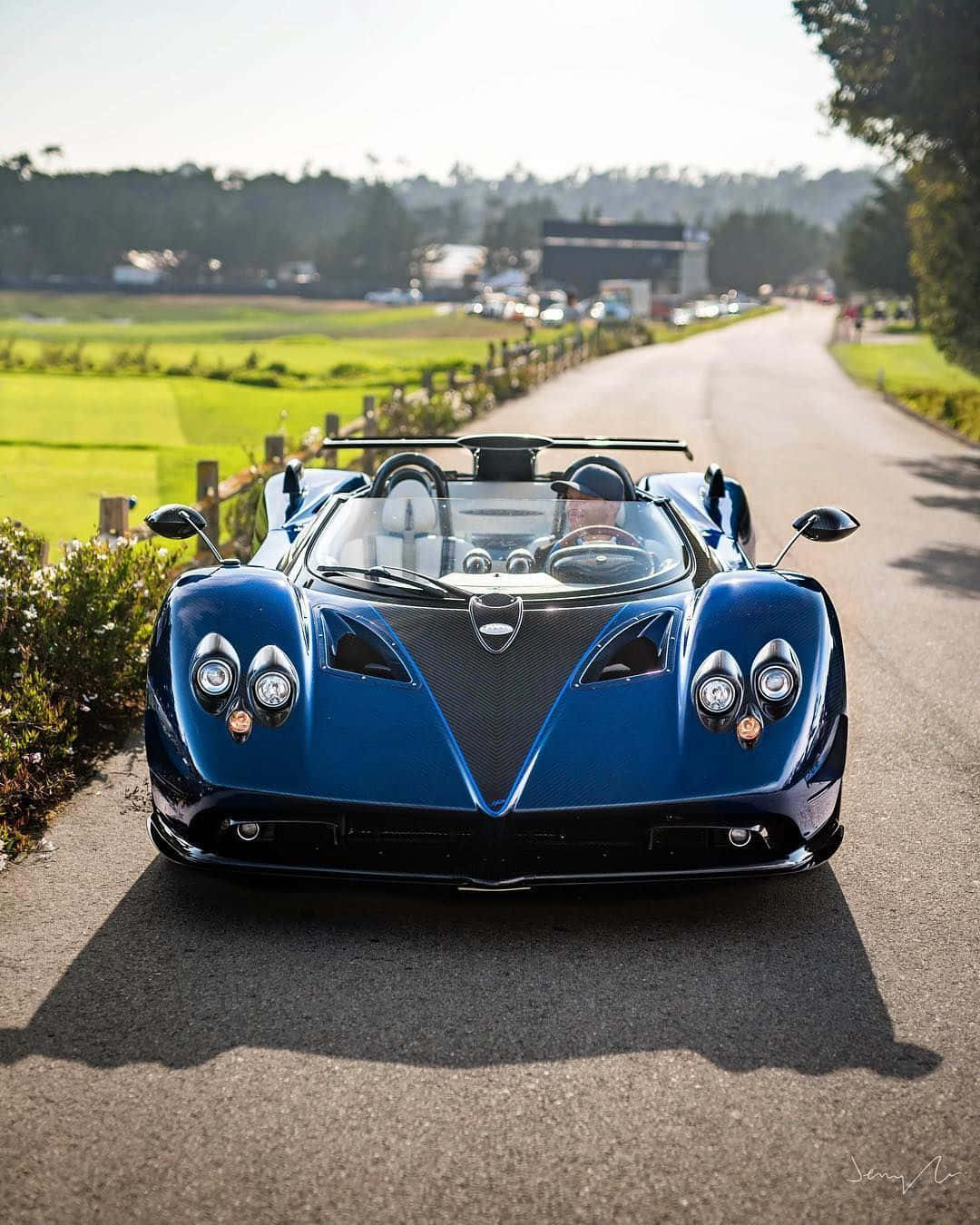 Pagani Zonda Hp Barchetta 桌布 壁紙 — 一輛時尚的藍色Pagani Huayra開篷跑車停泊在陽光明媚的鄉村道路上，背景是綠色的田野和樹木。
