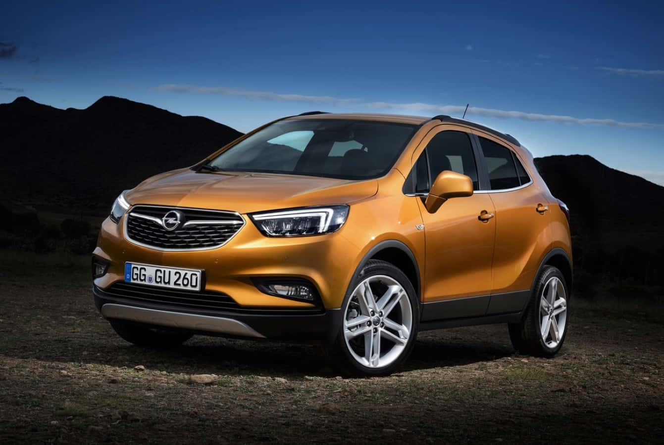 Opel Mokka 桌布 壁紙 — 一輛鮮橙色的Opel Mokka SUV停泊在崎嶇地形上，深色山脈在清澈藍天下。