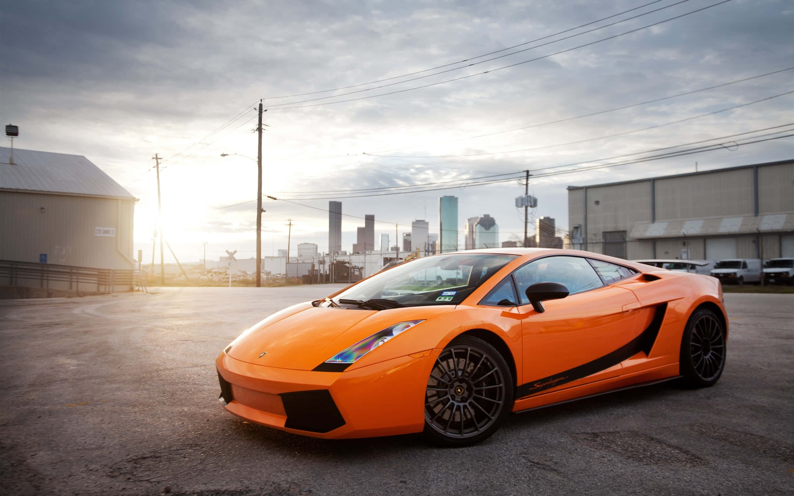 Lamborghini Gallardo 桌布 壁紙 — 一輛鮮橙色的林寶堅尼 Gallardo 停泊在工業區，背景是夕陽下閃耀的城市天際線。