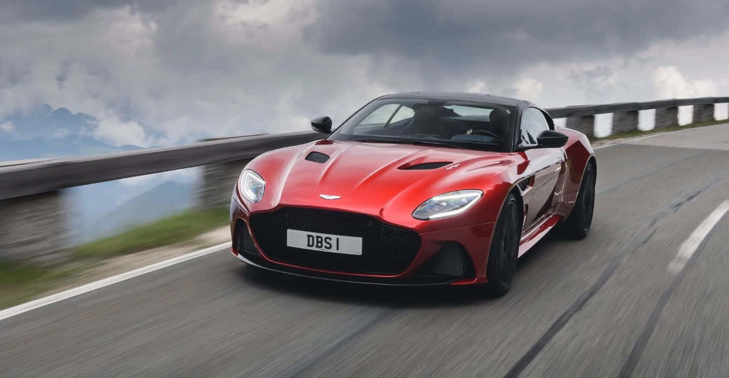 Aston Martin Dbs Superleggera 桌布 壁紙 — 一輛鮮紅色的Aston Martin DBS Superleggera在陰天之下沿著山路飛馳。