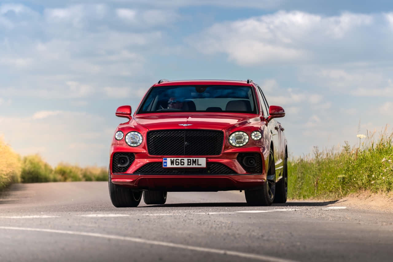 Bentley Bentayga 桌布 壁紙 — 一輛鮮紅色的賓利Bentayga SUV在部分多雲的天空下自信地行駛在鄉村道路上。