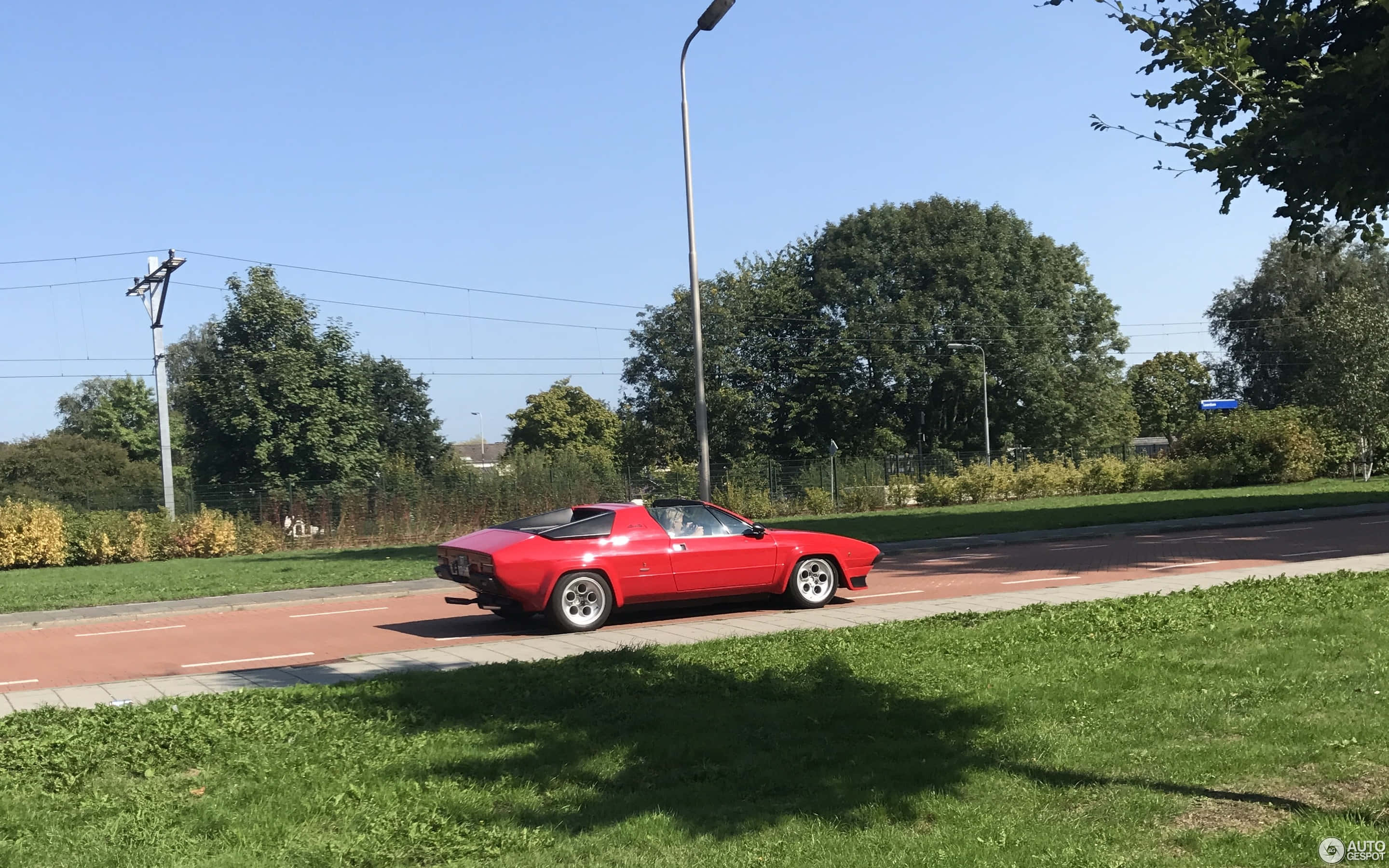 Lamborghini Silhouette 桌布 壁紙 — 一輛鮮紅色的經典跑車在晴朗的藍天下沿著綠樹成蔭的道路行駛。