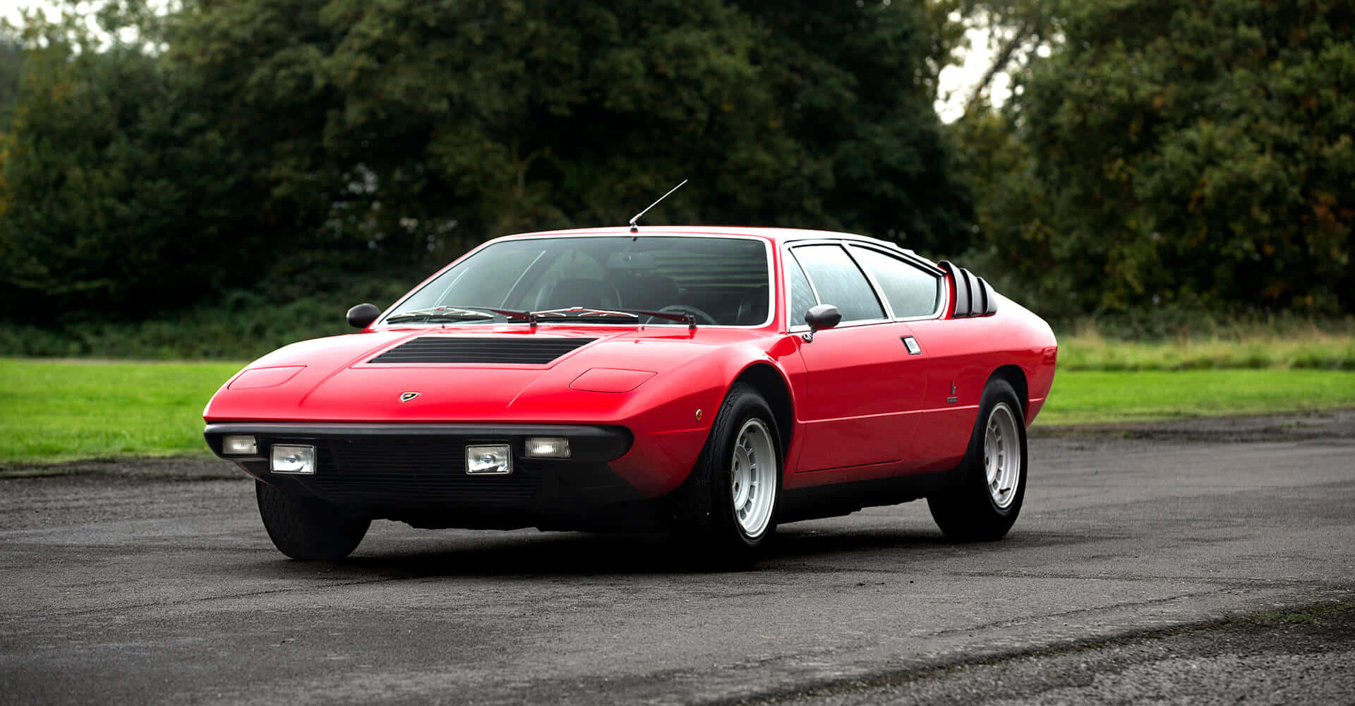 Lamborghini Urraco 桌布 壁紙 — 一輛鮮紅色的經典跑車，線條流暢，帶有黑色飾邊，停在鋪砌的道路上，周圍環繞著茂密的綠樹。