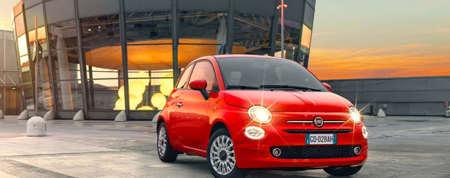 Fiat 500 桌布 壁紙 — 一輛鮮紅色的菲亞特500在溫暖的日落時分停在一棟現代玻璃建築前。
