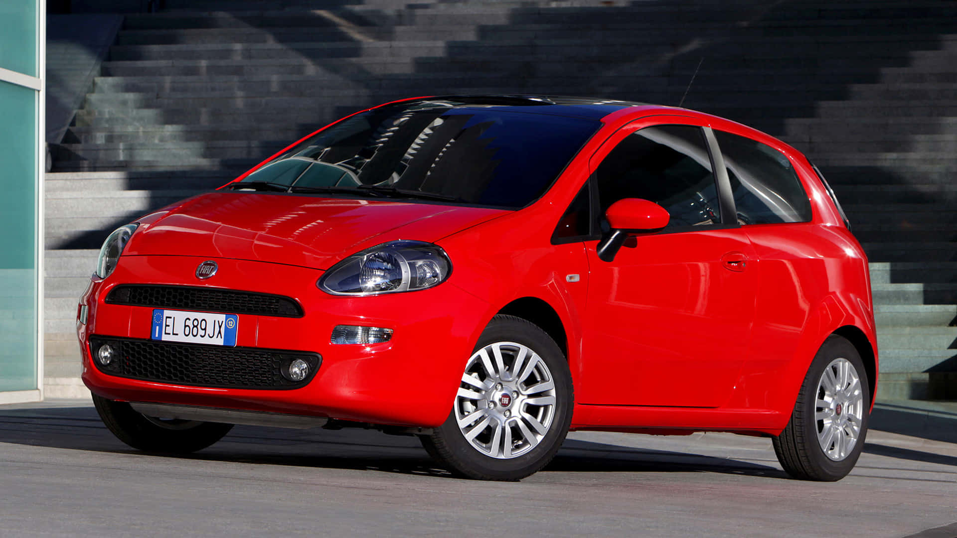 Fiat Grande Punto 桌布 壁紙 — 一輛鮮紅色的菲亞特Punto在明亮的日光下停泊在現代混凝土樓梯旁的行人路上。