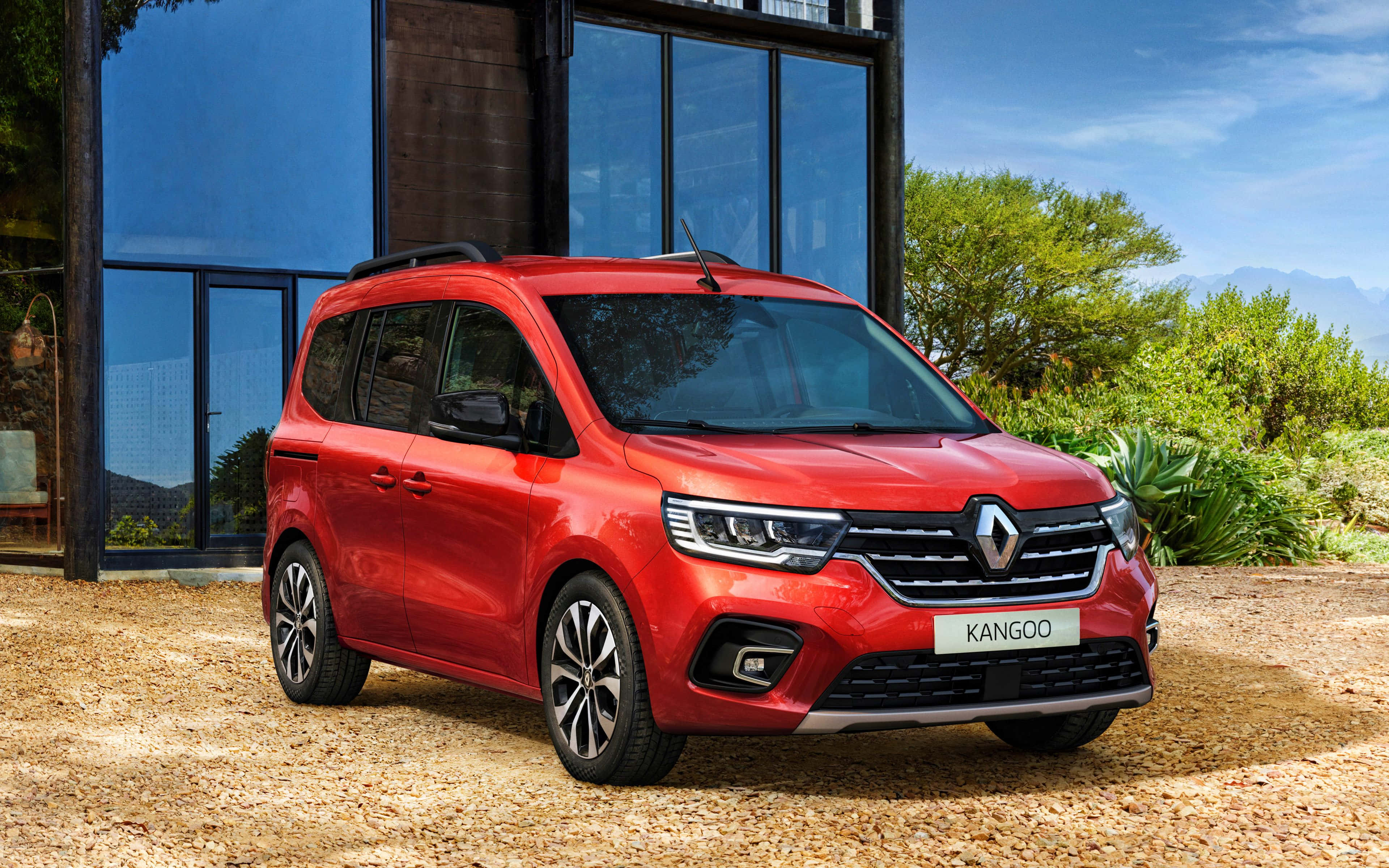 Renault Kangoo 桌布 壁紙 — 一輛鮮紅色的雷諾Kangoo客貨車停在碎石上，前方是一座現代玻璃屋，周圍有茂密的綠植和遠處的山脈，在清澈的藍天下。