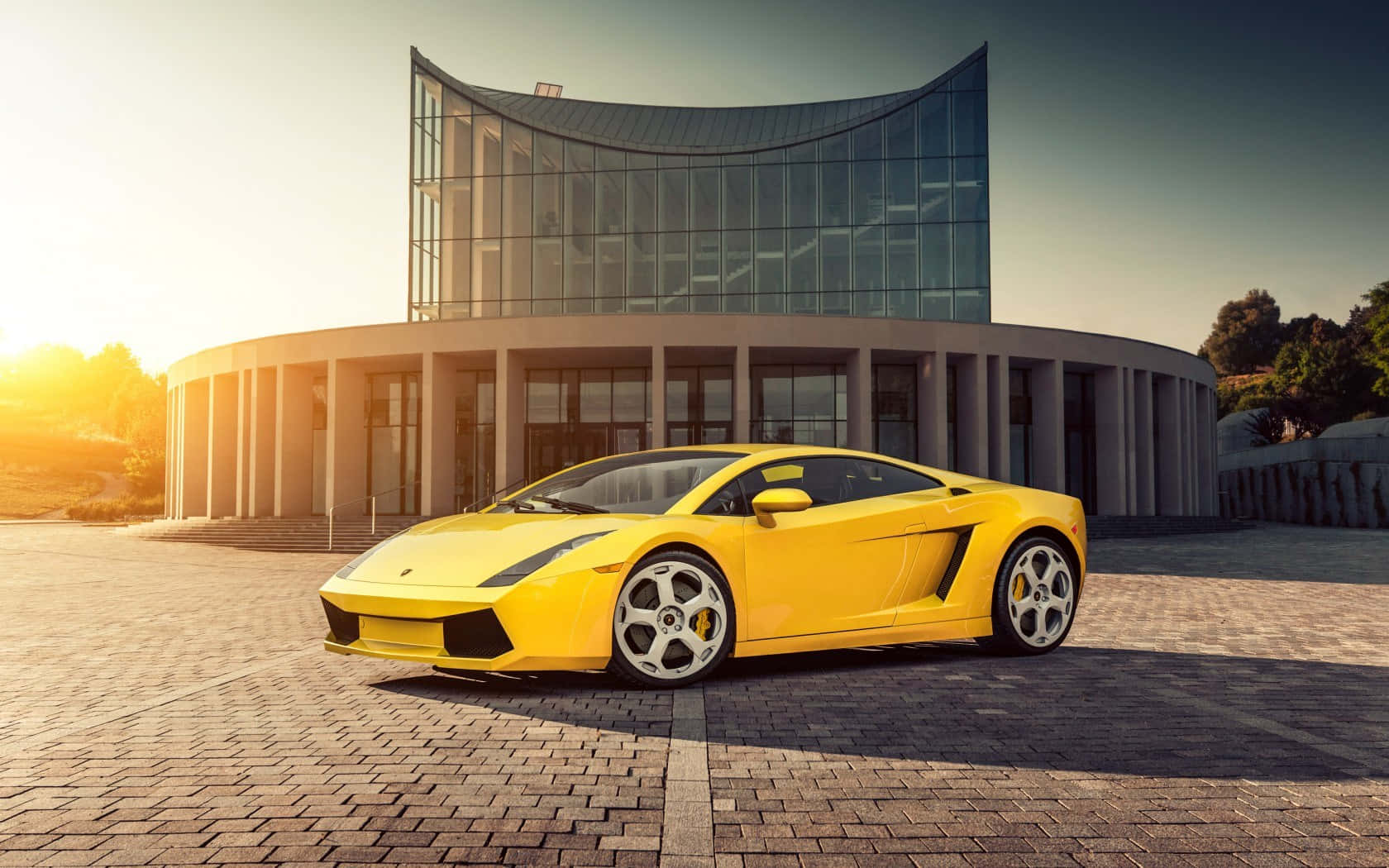 Lamborghini Gallardo 桌布 壁紙 — 一輛鮮黃色的林寶堅尼Gallardo停泊在現代玻璃建築前陽光普照的磚砌廣場上。