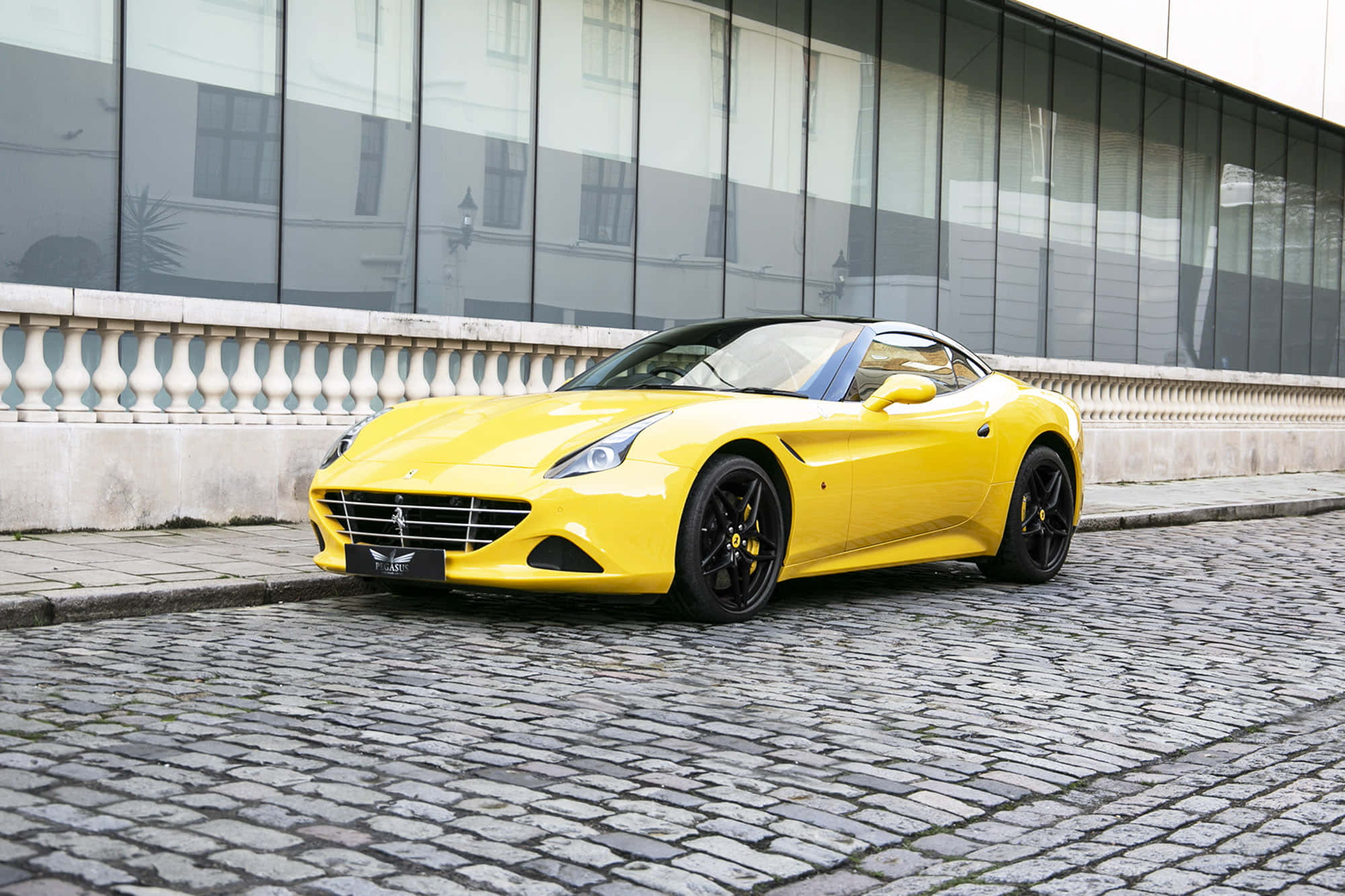 Ferrari California T 桌布 壁紙 — 一輛鮮黃色的法拉利跑車停泊在現代玻璃建築旁的鵝卵石街道上。