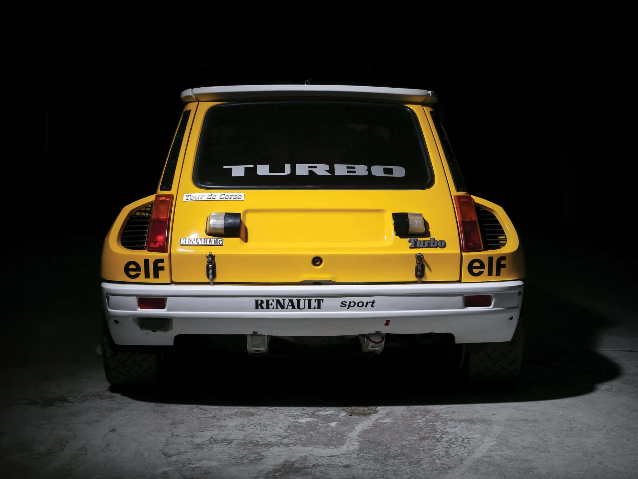 Renault 5 Turbo 桌布 壁紙 — 一輛鮮黃色雷諾5 Turbo跑車的尾部在黑暗背景下被照亮，展示其標誌性賽車貼紙和侵略性設計。