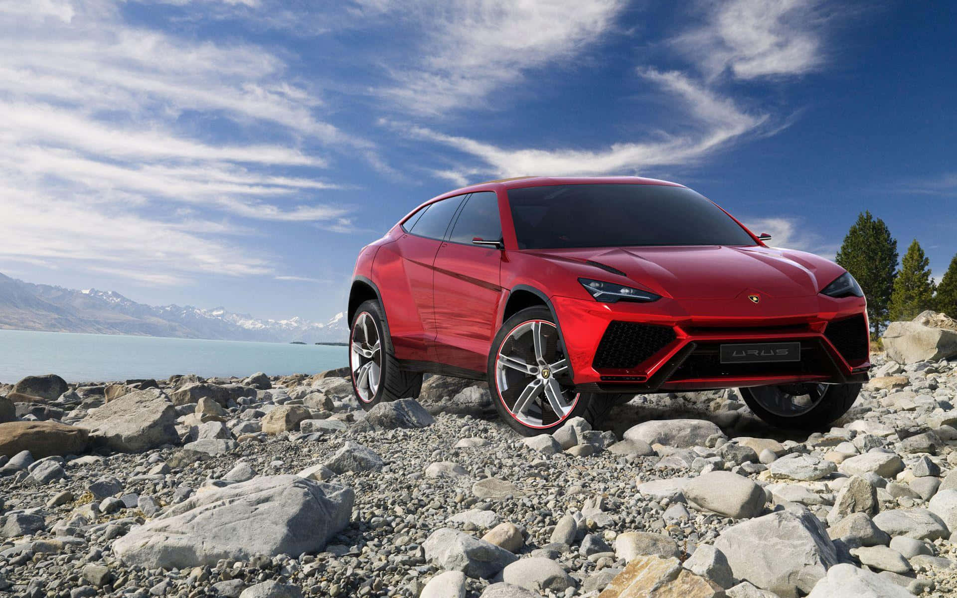 Lamborghini Urus 桌布 壁紙 — 一輛醒目的紅色林寶堅尼 Urus SUV 大膽地停在岩石湖岸上，背景是白雪覆蓋的山脈和明亮的藍天。