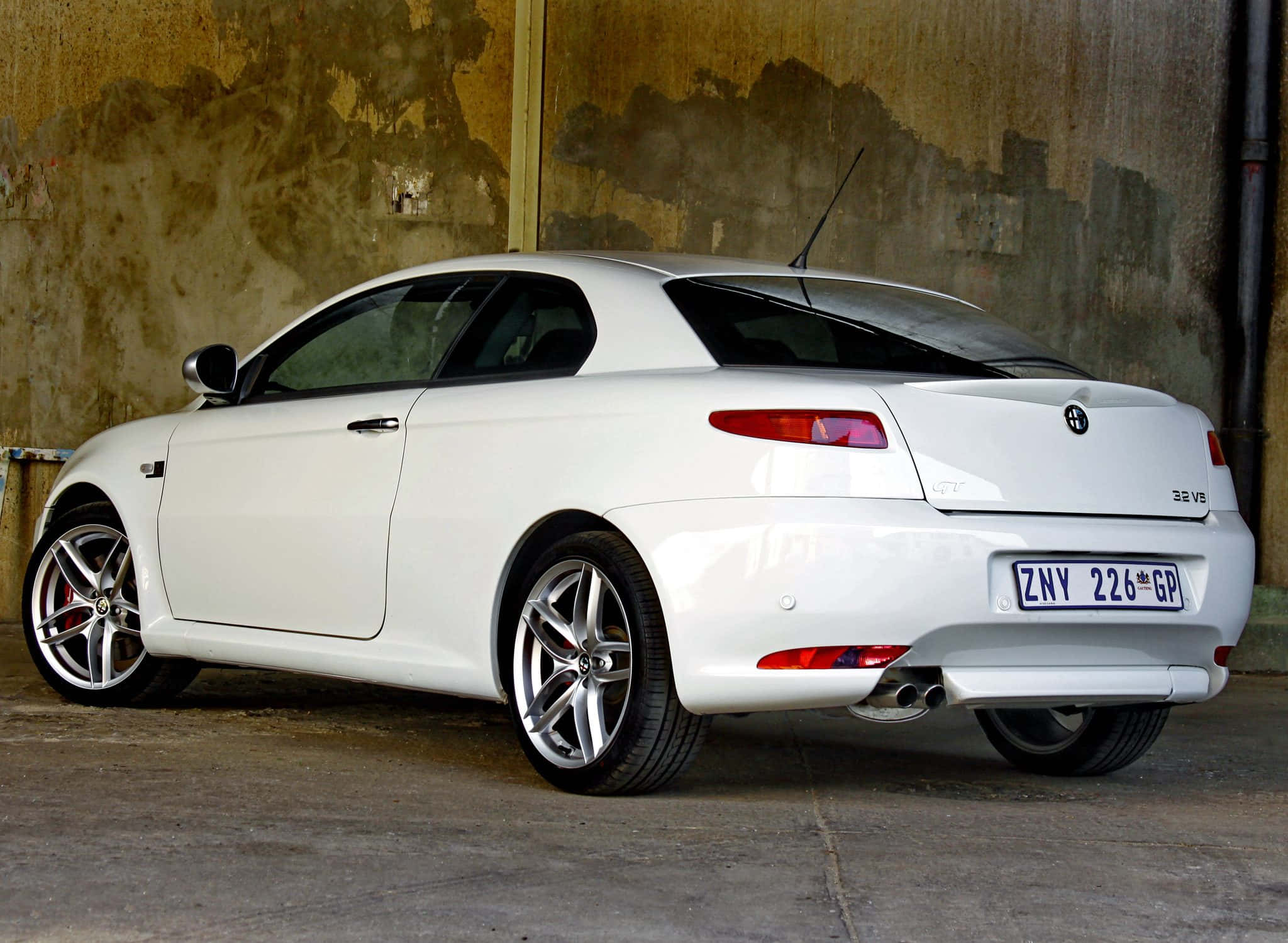 Alfa Romeo Gt 桌布 壁紙 — 一輛純白色的愛快羅密歐GT轎跑車停泊在光線昏暗的車庫中，倚靠著風化的混凝土牆。