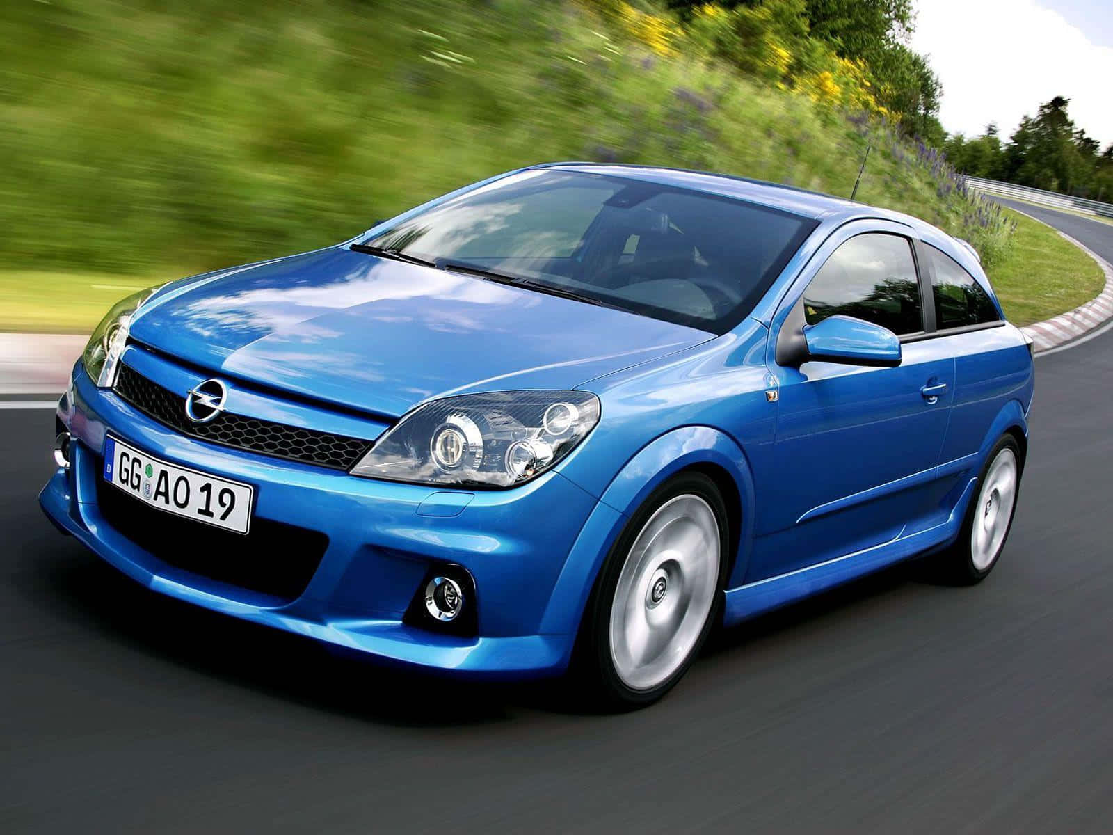 Opel Astra 桌布 壁紙 — 一輛充滿活力的藍色歐寶雅特GTC在彎曲的道路上疾馳，背景是模糊的綠色植物和野花。