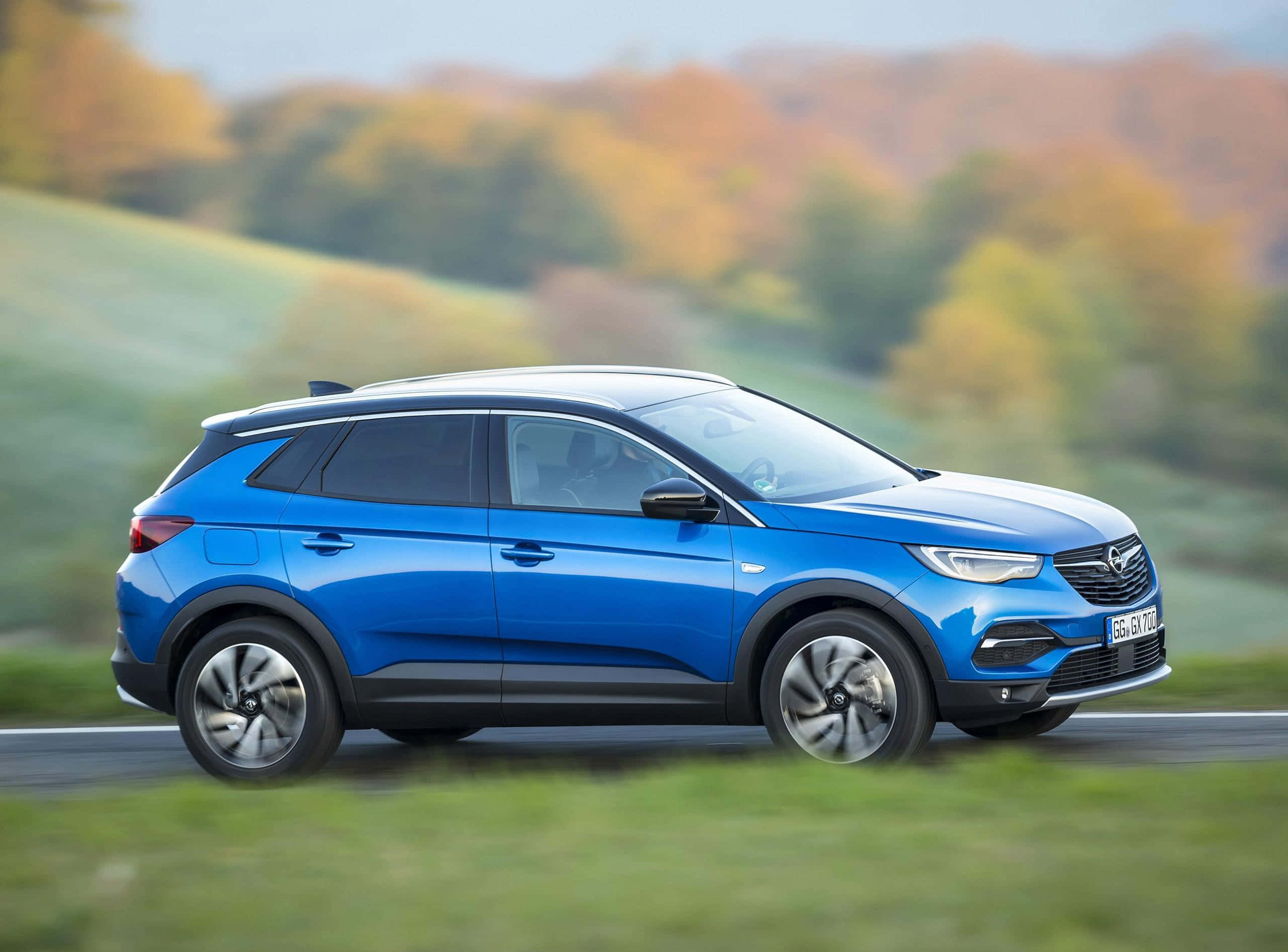 Opel Grandland X 桌布 壁紙 — 一輛充滿活力的藍色SUV沿著蜿蜒的道路行駛，穿過色彩繽紛的秋日景觀，樹葉模糊。