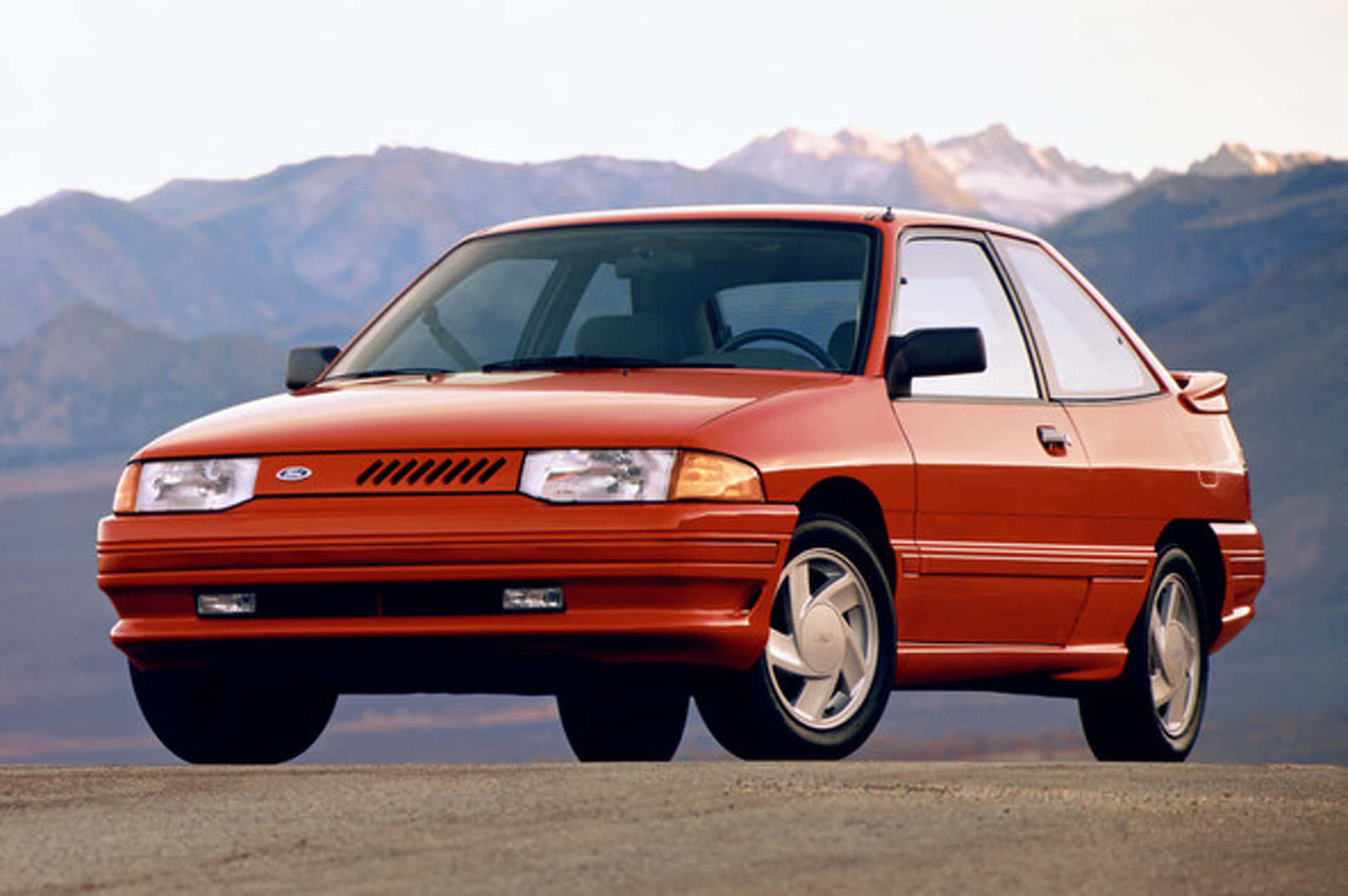 Ford Escort 桌布 壁紙 — 一輛鮮橙色的福特Escort雙門轎跑車停在沙漠公路上，背景是積雪的山脈。