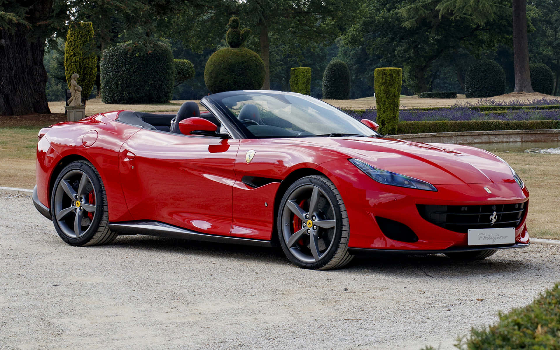 Ferrari Portofino 桌布 壁紙 — 一輛鮮紅色的法拉利 Portofino 開篷車在日光下閃耀，行駛在由修剪整齊的花園和雕塑式樹籬環繞的碎石路上。