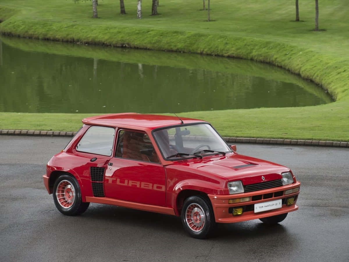 Renault 5 Turbo 桌布 壁紙 — 一輛鮮紅色的雷諾5 Turbo跑車停泊在潮濕的路面上，旁邊是一個寧靜的綠色高爾夫球場池塘。