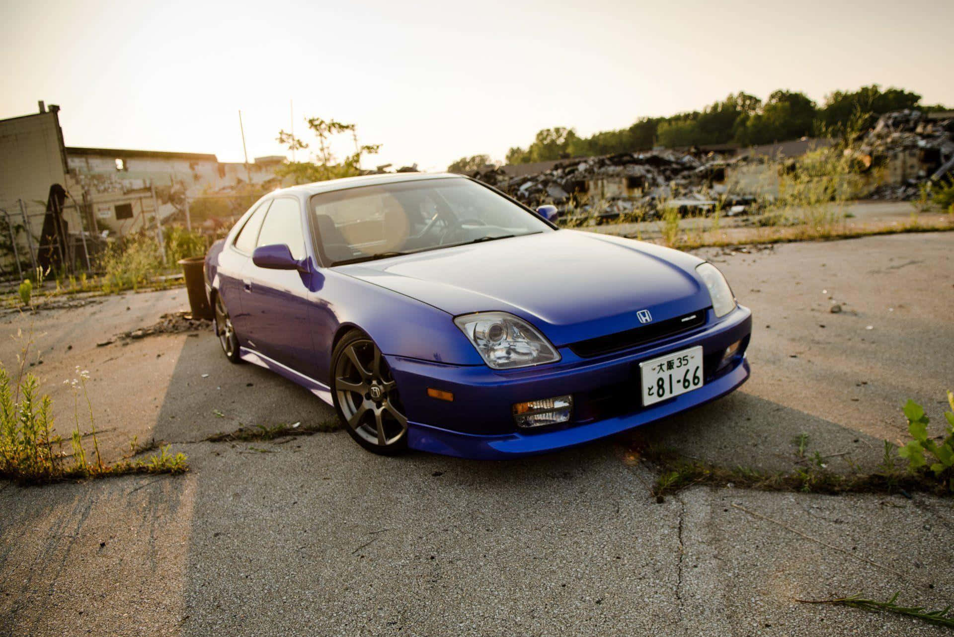Honda Prelude 桌布 壁紙 — 一輛鮮藍色的本田轎跑車停在裂開的瀝青路上，夕陽照射下，長長的影子投射在雜草叢生和廢棄建築物上。
