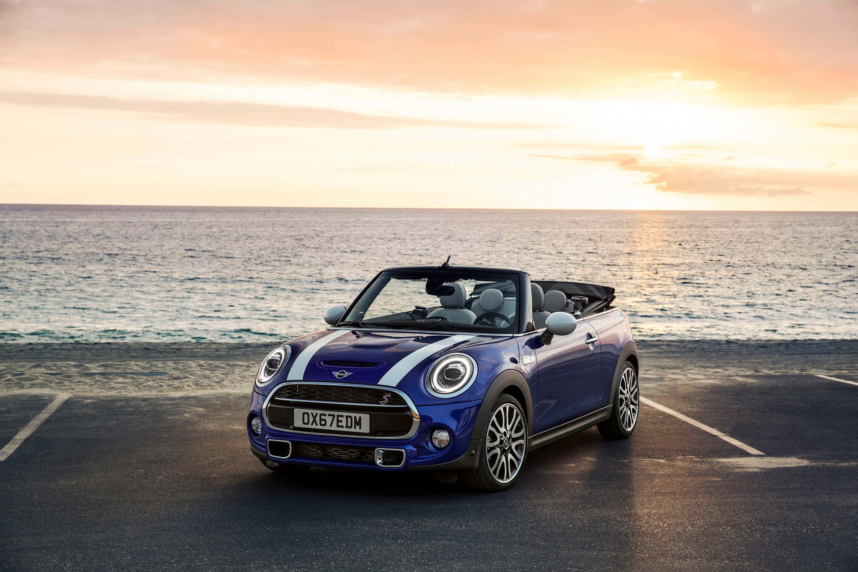 Mini Cooper Convertible 桌布 壁紙 — 一輛鮮藍色的MINI開篷車停在日落時分的海邊道路上，海浪翻滾而來。