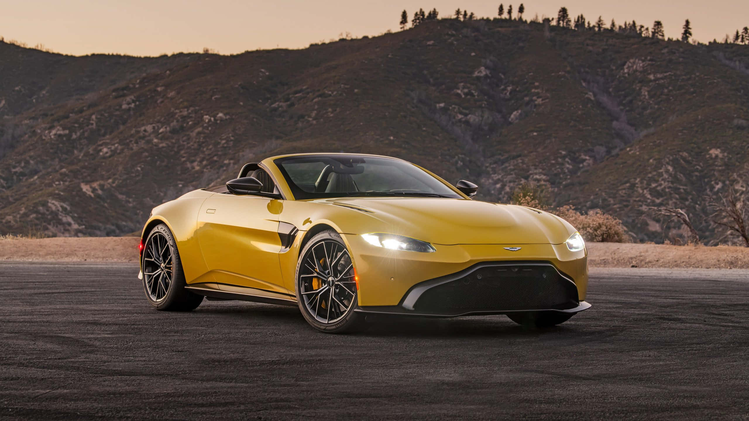 Aston Martin Vantage 桌布 壁紙 — 一輛鮮黃色的Aston Martin Vantage開篷車在陽光照耀的山脈背景下，在瀝青路上閃閃發光。