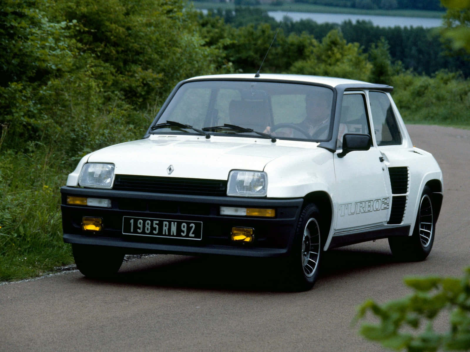 Renault 5 Turbo 桌布 壁紙 — 一輛白色1985年雷諾5 Turbo2在蜿蜒的道路上行駛，周圍環繞著茂密的綠樹和遠處的湖泊。