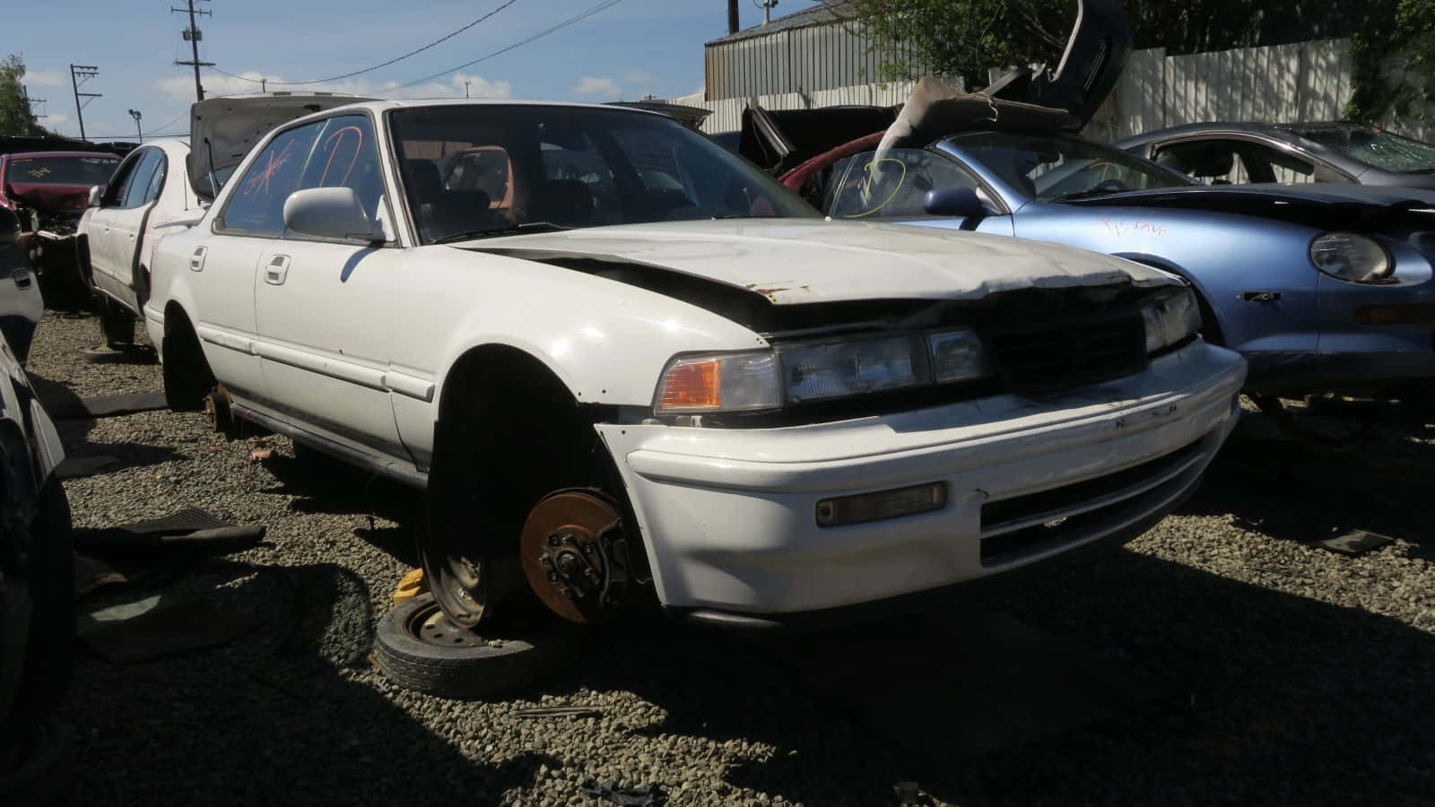  壁紙 — 一輛白色轎車，引擎蓋受損且缺少一個輪胎，被遺棄在明亮的陽光下，與其他報廢車輛一起停在碎石場。